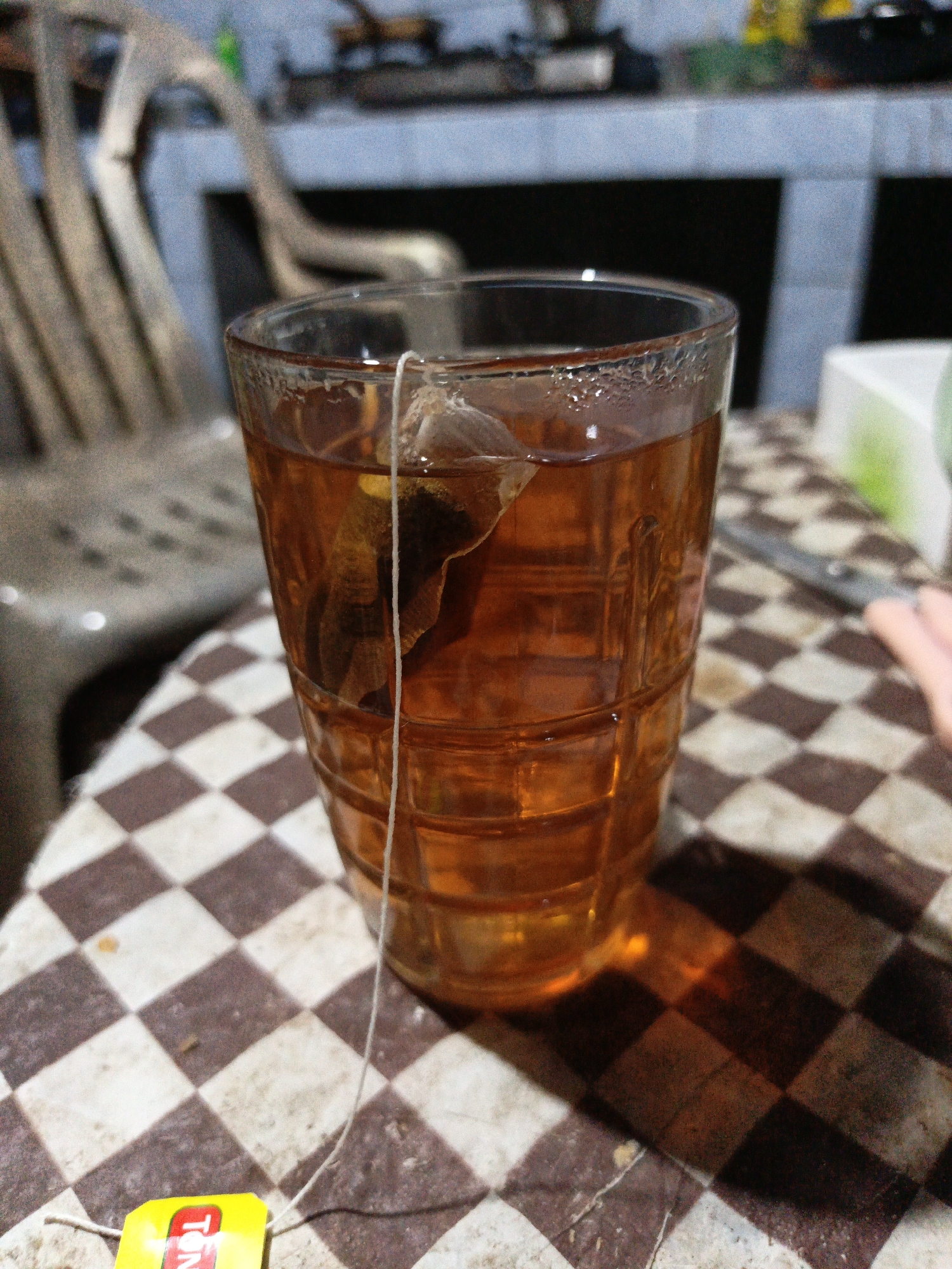 A close-up shot focuses on a glass of amber-colored tea with a tea bag steeping inside. The glass has a ribbed texture and is filled almost to the brim. A white string from the tea bag dangles down, with a yellow tag visible at the bottom, displaying red lettering that reads "TON." The glass sits on a checkered surface with alternating brown and white diamond patterns. In the background, out of focus, a light-colored chair with vertical slats and a darker structure resembling a stove or counter are visible. The lighting suggests an indoor setting, possibly a kitchen or dining area, with shadows cast on the checkered surface. The image appears to be taken in Gedung Nyapah, Indonesia, given the context of the question.