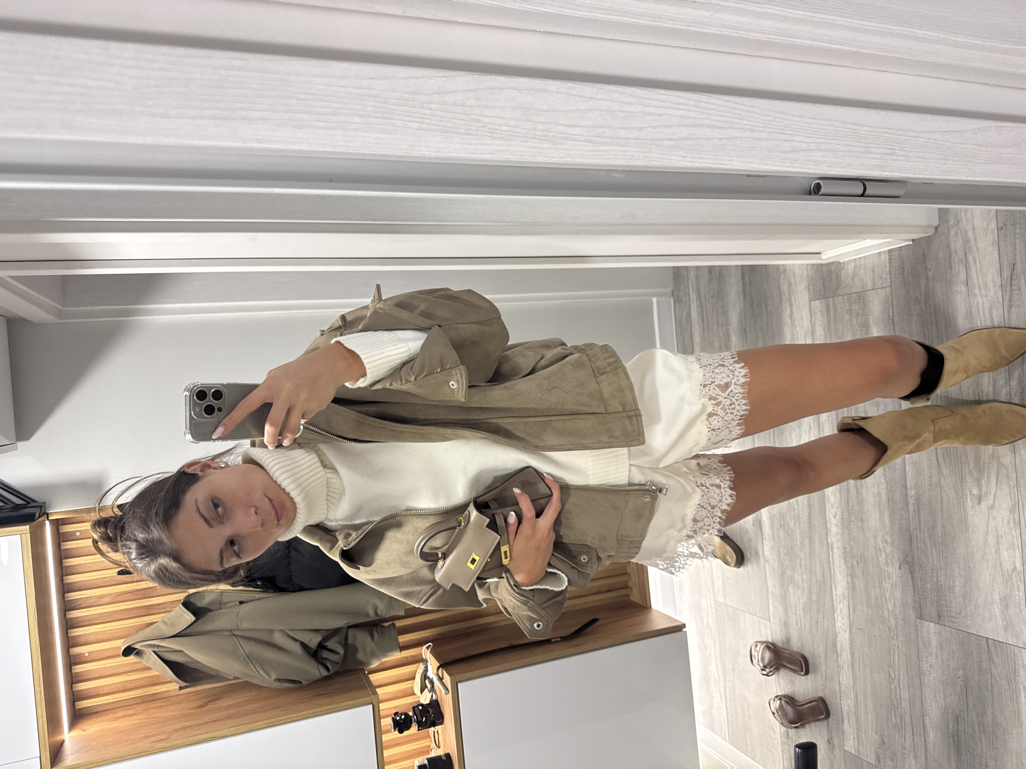 A young woman is taking a mirror selfie in what appears to be a hallway or dressing area. She is wearing a white turtleneck sweater, a beige suede jacket, a white lace-trimmed dress, and tan suede cowboy boots. She is holding a small beige handbag. The background consists of a mirrored wall, a wooden paneled wall with lighting, and a grey wood-effect floor. There are two pairs of sandals on the floor. The scene is well-lit, suggesting daytime. No text is visible in the image.