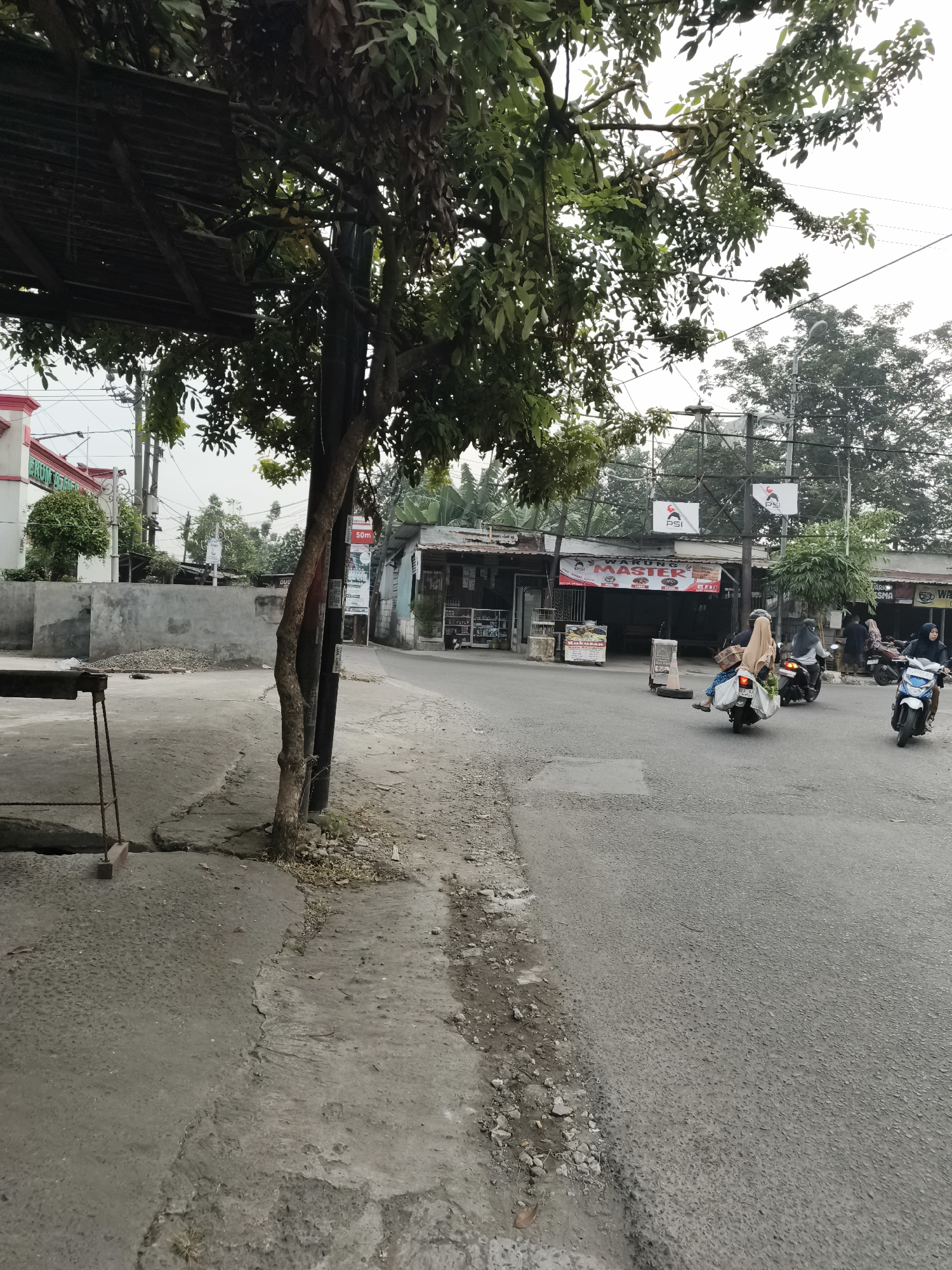 The image depicts a street scene in Sidorejo, Indonesia, likely during daylight hours with overcast skies. A narrow asphalt road dominates the right side of the frame, with a concrete sidewalk and a dirt shoulder on the left. Several motorcycles are present on the road; two are moving away from the viewer, and one is stationary. One motorcycle carries a passenger wearing a hijab.
Along the left side of the road, there is a raised concrete barrier and a tree with dense green foliage. A dark, rusted metal structure with a corrugated roof is partially visible on the far left. Behind the barrier, a low concrete wall is visible, with a pile of gravel in front of it. Further down the street, buildings with storefronts are visible, some with signage. One prominent sign reads "WARUNG MASTER". Power lines and poles are visible in the background, along with more trees and vegetation. The overall impression is of a typical urban or suburban street in Indonesia.