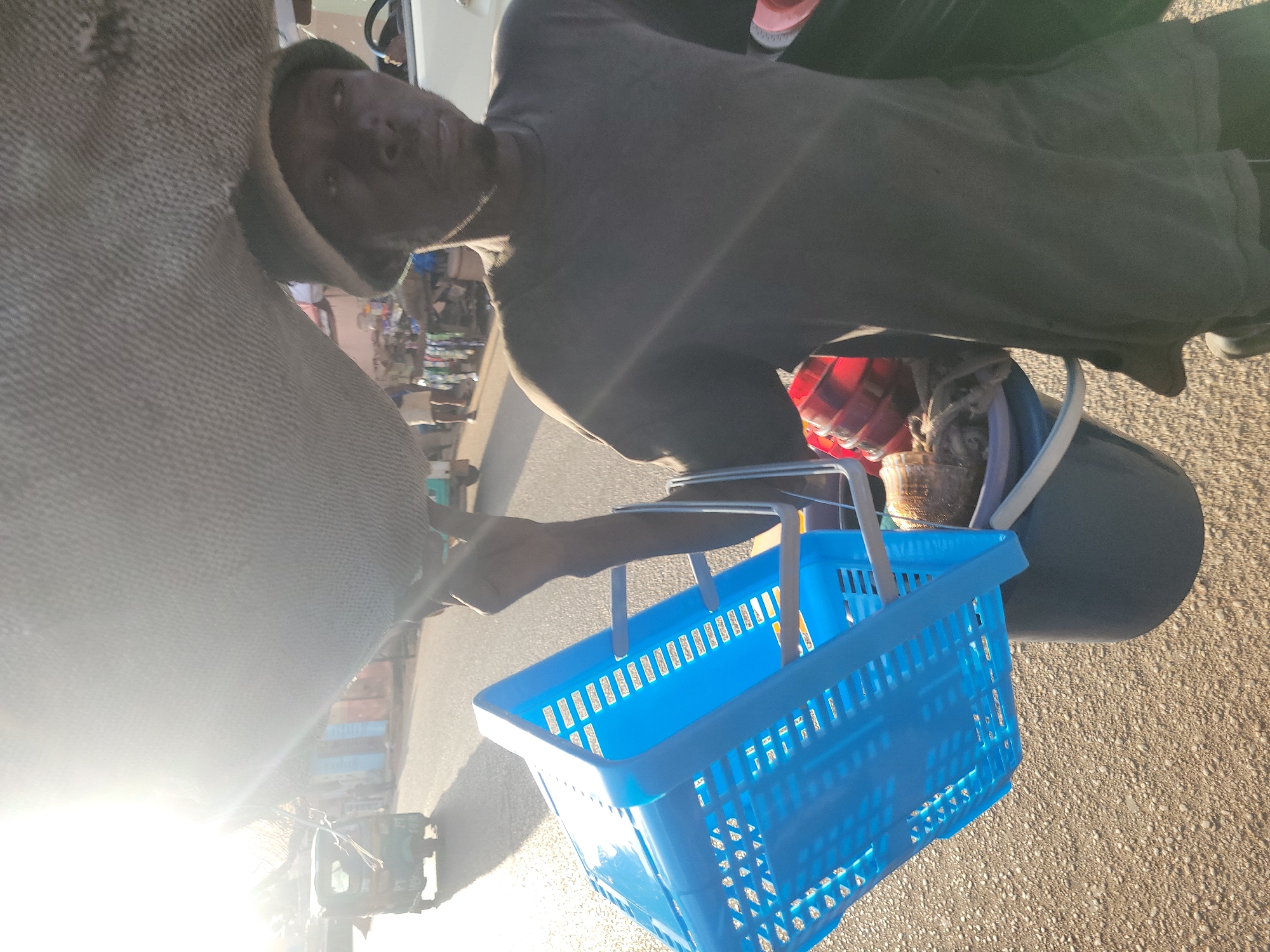 An individual, likely male, with dark skin, wearing a dark t-shirt and a dark, possibly woven, hat, walks along a paved road under bright sunlight in Jos, Nigeria. A very large, light-colored, irregularly shaped bundle, possibly fabric-wrapped, is balanced on the person's head, obscuring the upper right portion of the scene. In the individual's left hand, a bright blue rectangular plastic basket with handles is held, beneath which at least two dark-colored buckets are carried. One bucket contains various items, including multiple red, spherical, ribbed objects and other indistinct contents. The person's face is partially obscured by shadow due to strong backlighting from the upper left, where a significant sun flare is present. In the background, a three-wheeled auto-rickshaw is visible on the road, alongside various buildings with colored facades (blue, light brown) and street vendors or shops. A white vehicle is partially visible to the right of the individual. The road surface is light brown and textured.
