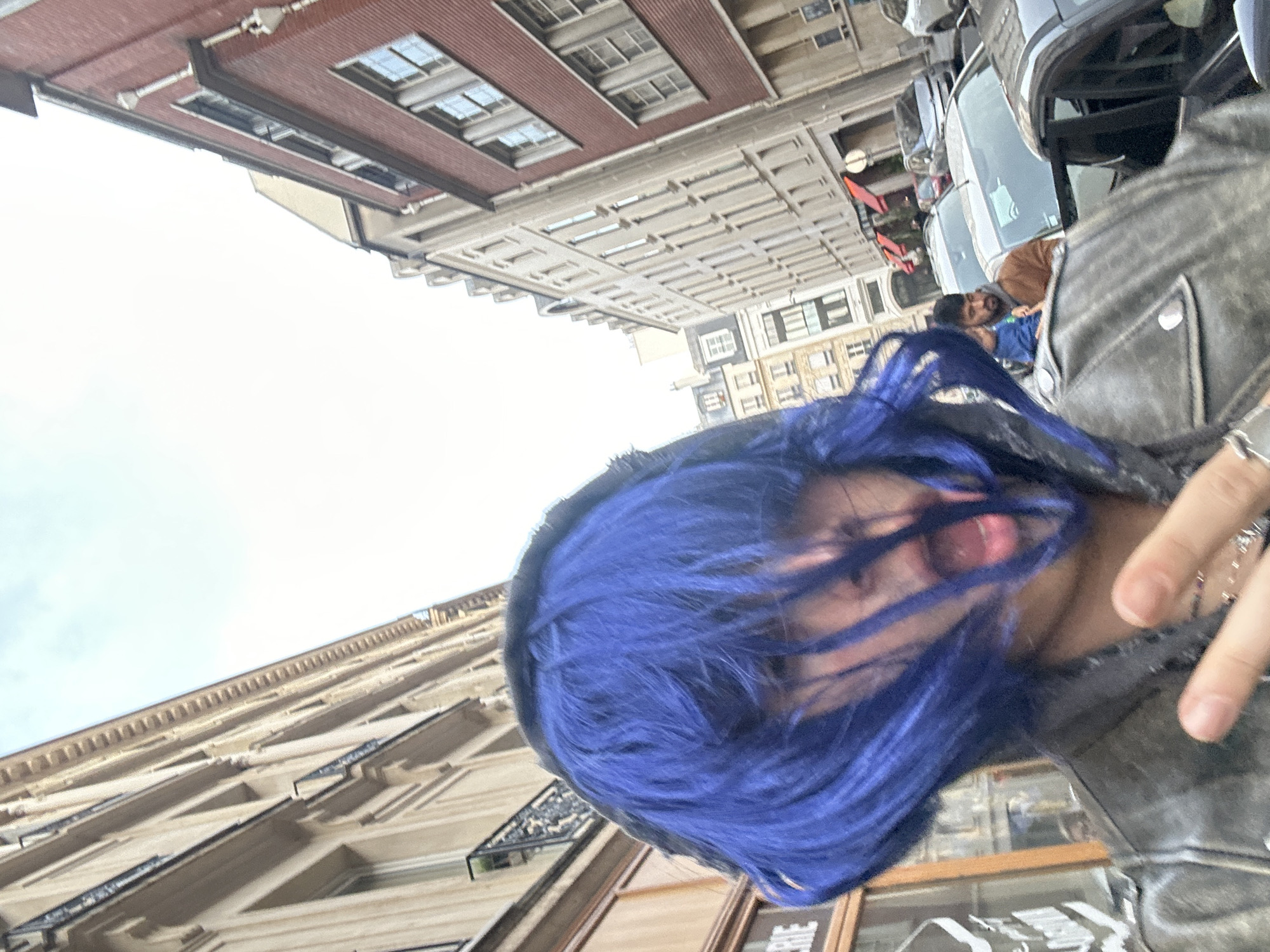 A first-person-perspective shot captures a person with blue hair and a gray leather jacket. They appear to be standing on a street in Paris, France. Behind them, tall buildings line the street. Several cars are parked along the side, and other people are visible further down the street.