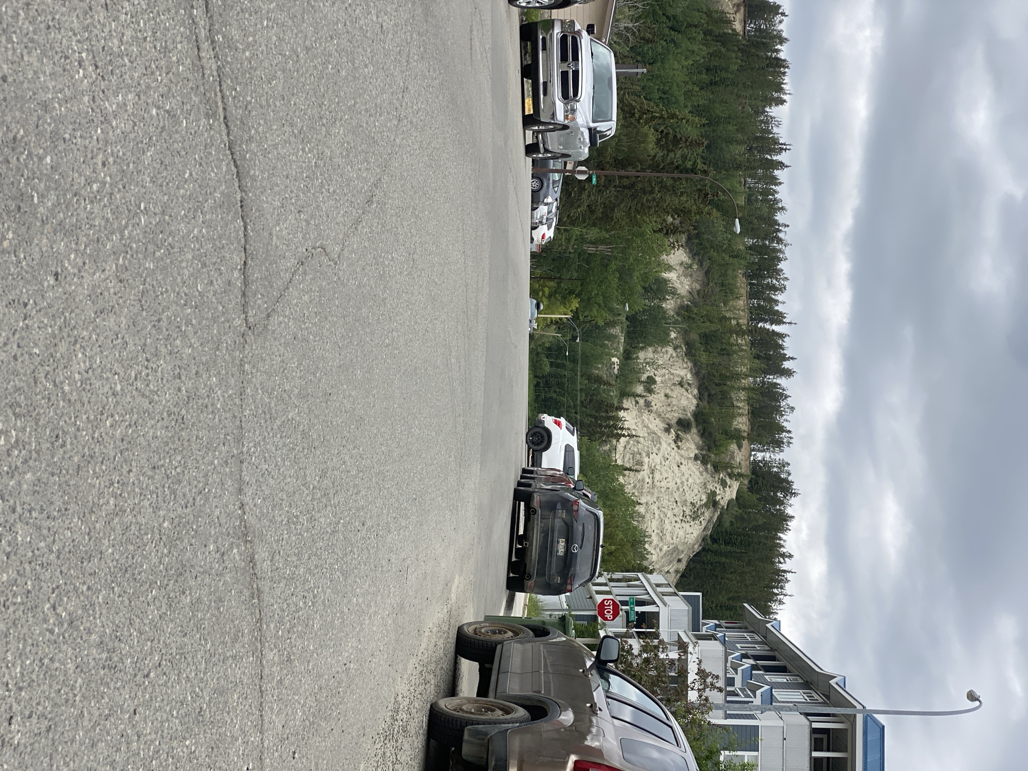The image shows a street scene in Whitehorse, Canada, on an overcast day. A line of parked cars is visible on the right side of the street, facing away from the viewer. The cars include a silver pickup truck, a dark SUV, a white SUV, and other vehicles further down the line.
Behind the cars, a steep, sandy embankment covered in evergreen trees rises upwards. The trees create a dense forest that extends to the top of the frame. The sky is filled with heavy, gray clouds, suggesting it might be a cloudy or rainy day.
In the foreground, the asphalt road is cracked in places. To the right of the road, a modern, multi-story building with a blue roof is visible. A street sign indicating "STOP" is mounted on a pole next to the building. There are no people visible in the image. The overall mood is calm and perhaps a bit subdued due to the overcast weather.