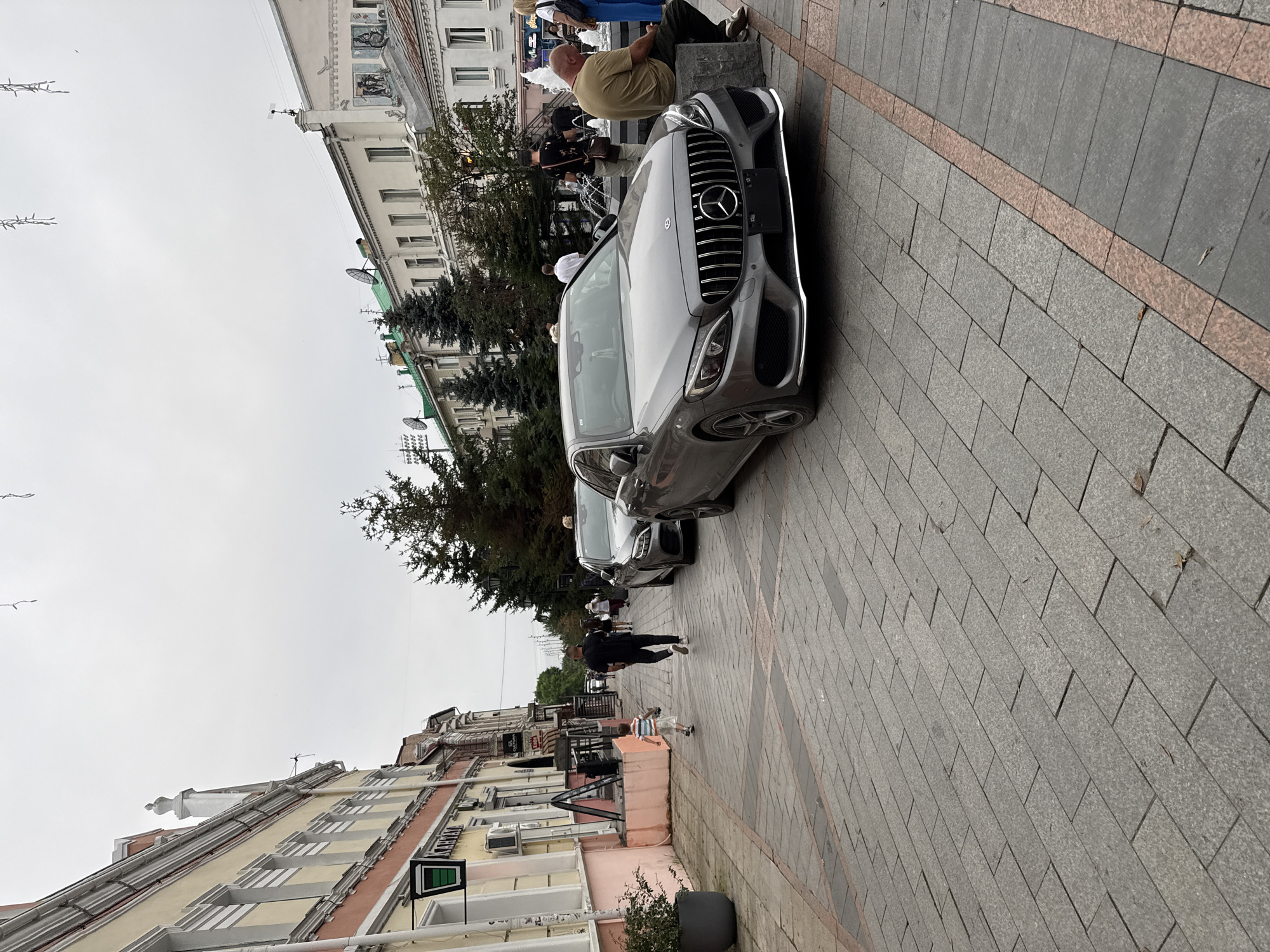 A street scene in Vladivostok, Russia, features a prominent gray Mercedes-Benz sedan parked on a pedestrian walkway paved with gray and reddish-brown interlocking stones. The car's grille, headlights, and front bumper are clearly visible. Several other cars, including a silver sedan, are parked behind it. A fountain with multiple tiers of water jets is visible in the background, as are several people walking and sitting. The buildings lining the street have light-colored facades with arched windows and decorative cornices. Trees are planted along the walkway. A young boy in a striped shirt and shorts is running on the walkway, and a man in a black t-shirt and pants is walking ahead of him. Another man wearing a light brown t-shirt is seated on a stone block near the fountain. The sky is overcast.