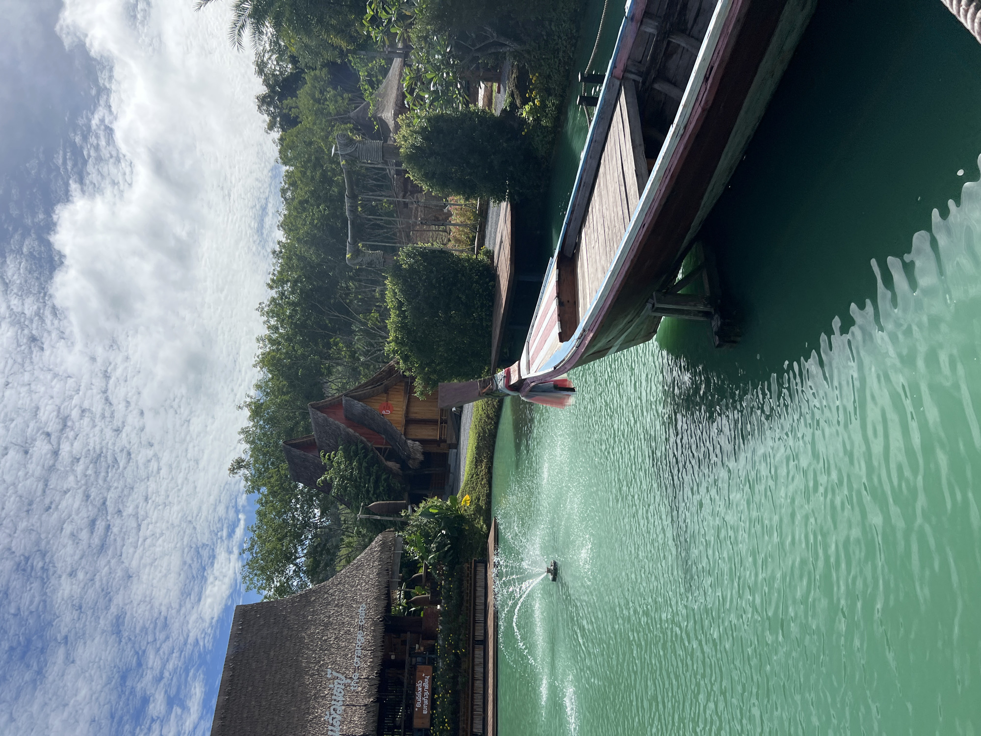An eye-level outdoor shot captures a tranquil scene in Tambon Pa Kae Bo Hin, Thailand. The water in a pond is a vibrant turquoise, disturbed by a small fountain in the left middle ground. A traditional longtail boat, painted with red, blue, and white accents, is docked at a wooden pier in the right foreground, its bow pointing away from the viewer. Behind the pond, thatched-roof buildings with wooden structures are visible, nestled amongst lush green trees. The sky above is a bright blue, dotted with fluffy white clouds. The overall atmosphere is peaceful and idyllic.