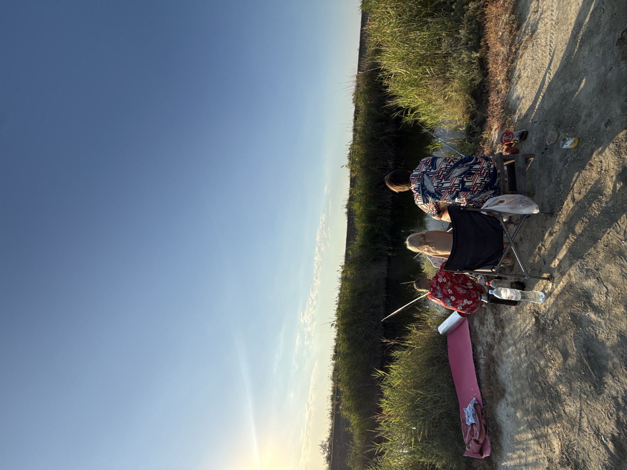 In the city of Некрасовское сельское поселение, Ukraine, two individuals, likely a child and an adult, are fishing by a body of water. The adult, wearing a patterned shirt, is seated in a folding chair. The child, in a red shirt, is positioned slightly in front of them. Both are facing the water, with fishing rods in hand, suggesting they are actively engaged in fishing. The environment is characterized by tall grass and reeds lining the water's edge, with a clear blue sky overhead. The sun is setting, casting a warm glow on the scene. A pink mat and a water bottle are visible on the ground, indicating a casual outdoor activity. The overall atmosphere appears peaceful and recreational.