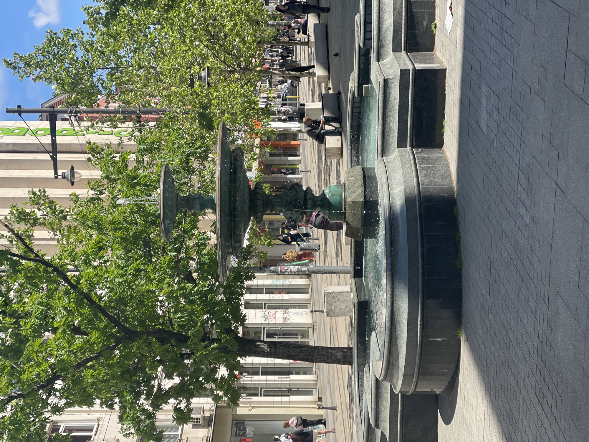 The image captures a street scene in Sofia, Bulgaria, featuring a large fountain. The fountain is multi-tiered, constructed of gray stone, and situated along a pedestrian walkway paved with square tiles. People are present in the scene, walking along the walkway, sitting on benches, or near the fountain. Trees with dense green foliage flank the fountain, adding shade to the area. Buildings line the street, displaying shop signs and graffiti. The sky is partly cloudy, suggesting a bright, sunny day.