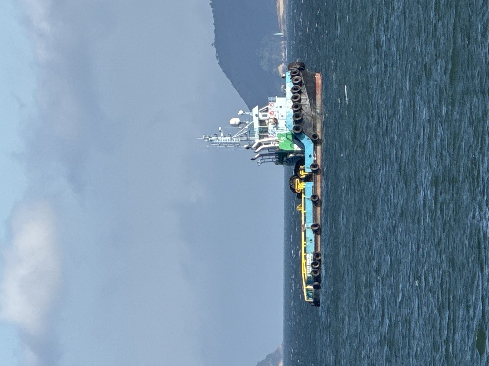 A tugboat, painted blue and green, is navigating through choppy, dark blue waters under a cloudy sky. The boat has a raised cabin structure with visible antennas and equipment. It is towing a flat barge, also painted blue with yellow railings, which is lined with black fenders. In the background, a hilly, green landmass is visible, possibly an island or coastline. The water is rough with visible waves. The scene appears to be in a coastal or harbor area, given the presence of boats and landforms. No people are clearly visible in the image.