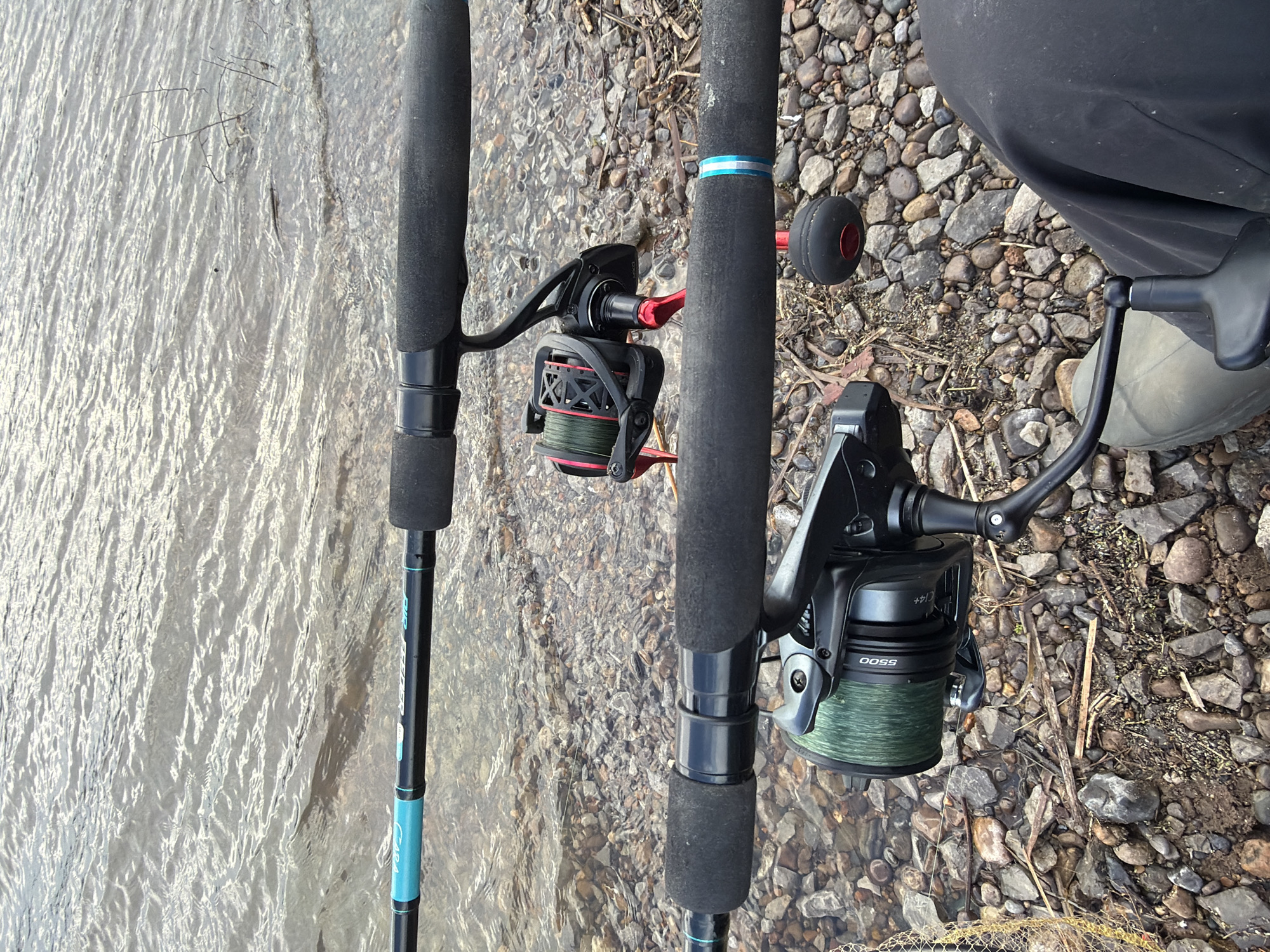 The image shows two fishing rods with reels, positioned on a rocky shore near water. The rod on the left is partially submerged in the water. The rod on the right is resting on the rocks. A person wearing dark pants and light-colored boots is partially visible on the right side of the frame. The rocks are a mix of sizes and colors, and the water has a reflective surface. There is no clear evidence indicating this photo was taken in the city of Chaikovsky, Russia.