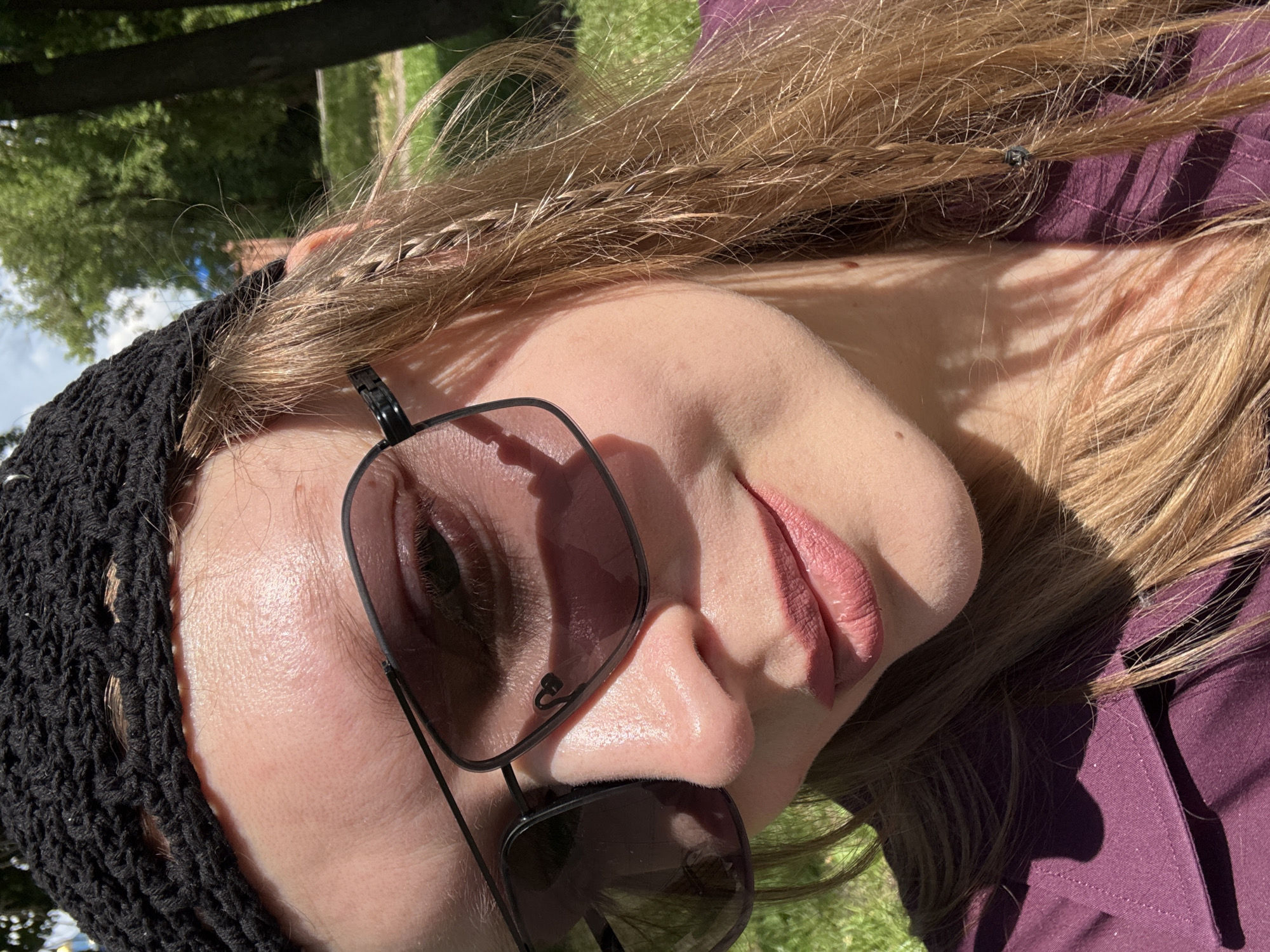 A close-up, overhead shot shows a person wearing a black crocheted hat and sunglasses, with their head tilted slightly to the left. The person has light skin and light brown hair, with a braid visible on the right side of their head. They are wearing a dark purple collared shirt. The background is blurred and appears to be an outdoor setting with green grass, trees, and a blue sky with some clouds. The lighting suggests it is daytime, possibly sunny with some dappled shade. The location is not specified as Izhevsk, Russia.