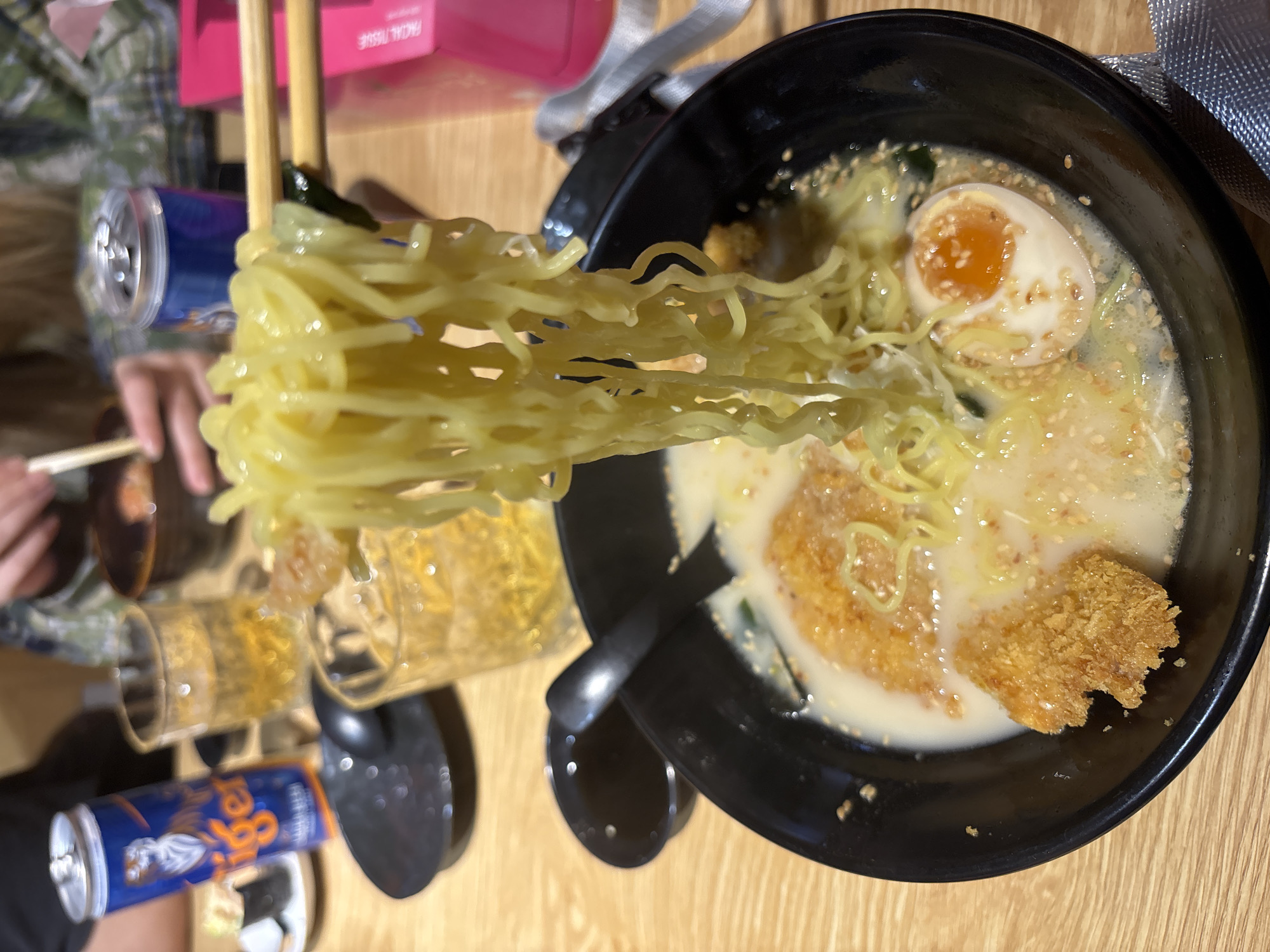 In Lâm Viên - Đà Lạt, Vietnam, a close-up shot captures a person holding chopsticks full of ramen noodles above a bowl of ramen. The ramen bowl contains a creamy broth, crispy fried chicken pieces, a soft-boiled egg cut in half, and sesame seeds. In the background, several glasses of amber liquid, a can of Tiger beer, and a pink tissue box are visible on a wooden table. The scene suggests a casual dining experience.