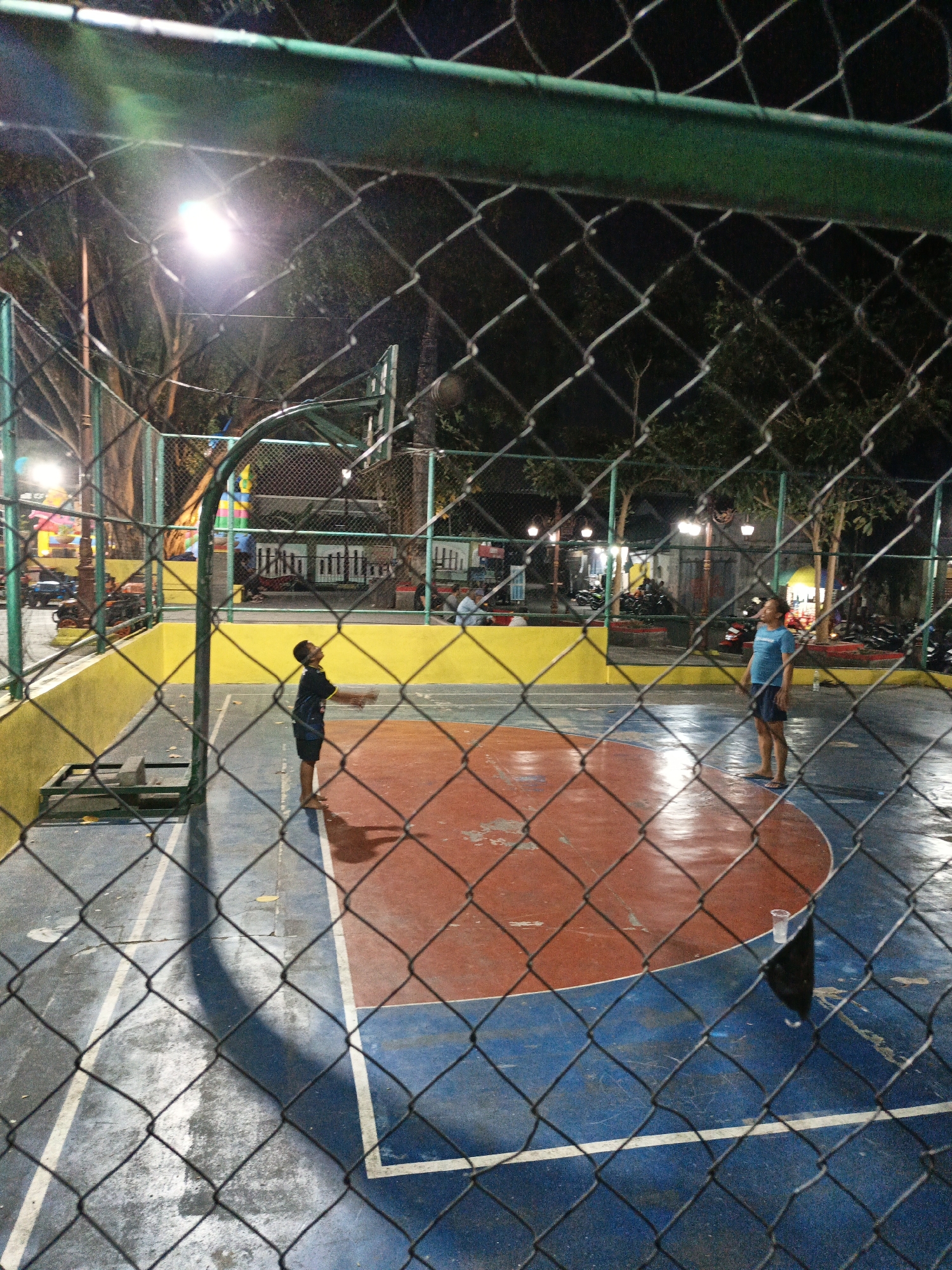 The image shows an outdoor basketball court at night, possibly in the city of Klaten, Indonesia. A chain-link fence runs across the foreground, partially obscuring the scene. The court has red and blue markings. Two boys are visible on the court: one on the left is wearing a dark blue shirt and shorts and appears to be looking up and raising his hands, possibly playing basketball. Another boy, wearing a blue t-shirt and dark shorts, stands on the right side of the court, facing away from the camera. In the background, there are trees, streetlights illuminating the court, and parked motorcycles. There are also some buildings and other people in the distance.