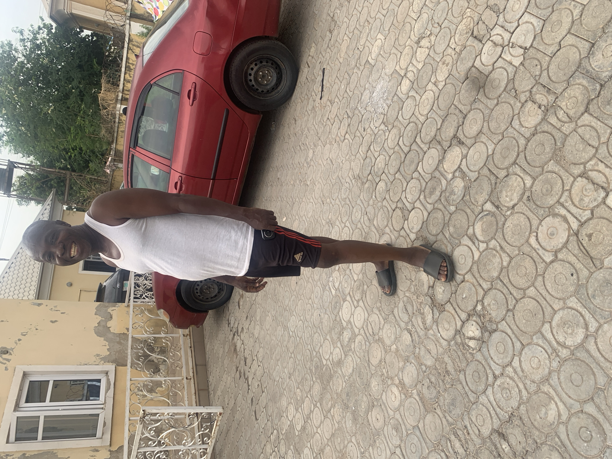 A man stands on a patterned concrete driveway in front of a faded yellow building in Abuja, Nigeria. The building has white-framed windows and decorative white metal railings. A red sedan is parked nearby. The man is wearing a white tank top, black shorts, and sandals. He smiles at the camera. A tree is visible in the background.