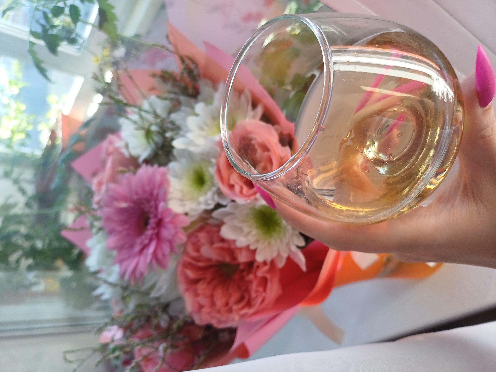 A hand with a bright pink stiletto nail holds a glass of pale yellow liquid, likely wine or champagne, in front of a blurred bouquet of flowers. The glass is clear and has some condensation on its surface. The bouquet is a mix of pink, white, and coral-colored flowers, including gerbera daisies and chrysanthemums, and is wrapped in clear cellophane with pink and orange ribbons. The background is out of focus, but suggests an indoor setting with a window, as evidenced by glimpses of greenery and natural light. The overall impression is one of celebration or relaxation, possibly a toast at a special occasion. There is no visible text.