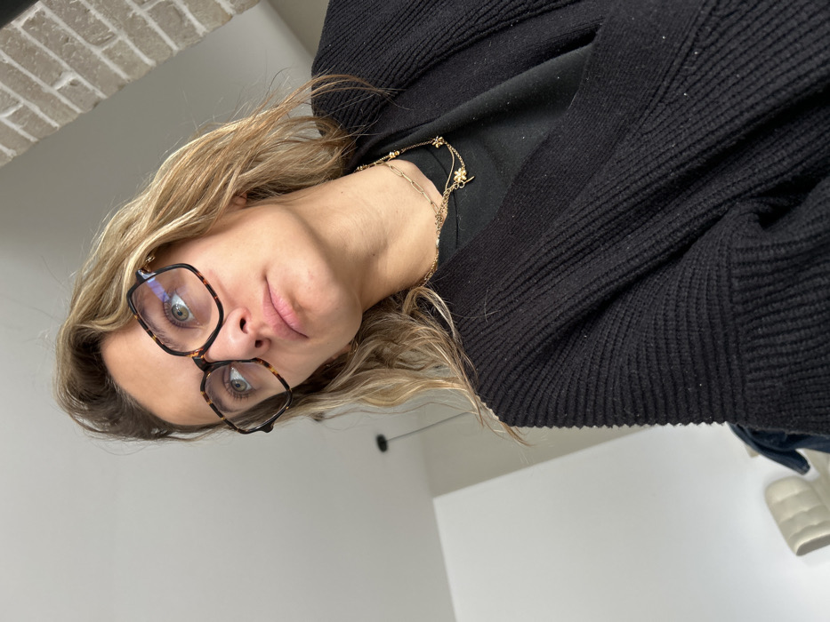 The image displays a close-up, low-angle perspective of an adult female positioned indoors in Veauchette, France. She has wavy, light brown hair and wears tortoise-shell patterned eyeglasses, through which her light-colored eyes are directed upwards and to the left. Her lips are slightly parted. She is dressed in a black, ribbed knitted cardigan over a plain black top, accessorized with layered gold-colored necklaces, one featuring multiple small flower or clover-shaped charms and a small key charm. The background elements include a segment of a light-colored brick wall on the upper right, a plain white wall or ceiling on the upper left, a dark suspended pendant light fixture visible mid-left, and a glimpse of light-colored, possibly upholstered, furniture in the lower left portion of the frame.