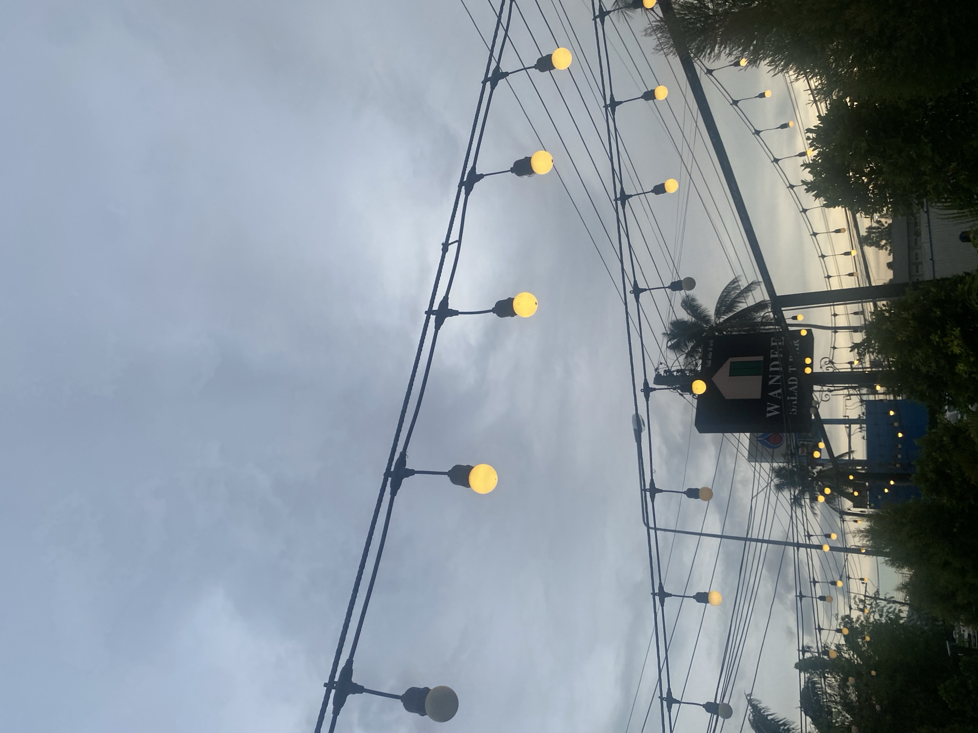 The image depicts an outdoor scene in Choeng Thale, Thailand, under an overcast sky. A string of lights with yellow bulbs stretches across the frame, intersecting with power lines. A sign for "WANDPEE SALAD TEP-PAK" is visible among trees and other lighting. The surroundings include foliage and glimpses of buildings.