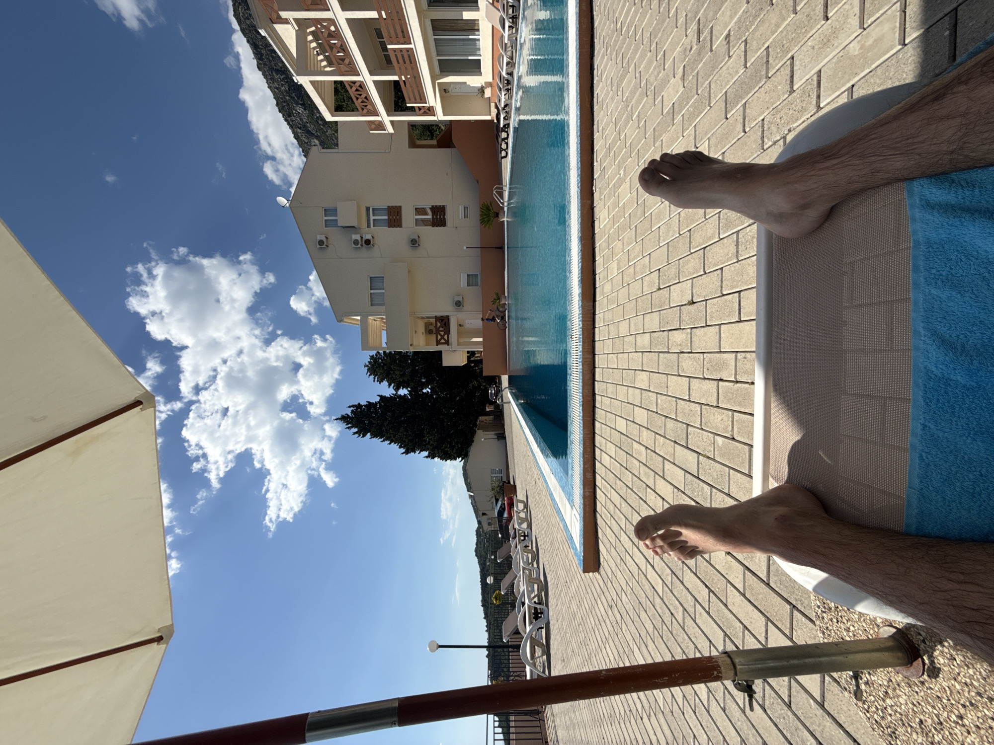 The image depicts a first-person view of someone relaxing on a lounge chair beside a swimming pool. The person's legs and feet are visible in the foreground, draped across a blue towel on the chair. The pool runs parallel to a brick-paved area. In the background, a building with multiple floors and balconies can be seen, along with trees and a mountain range. The sky is blue with scattered clouds. The location is identified as Šušanj, Montenegro, based on the visible architecture and landscape features of the image.