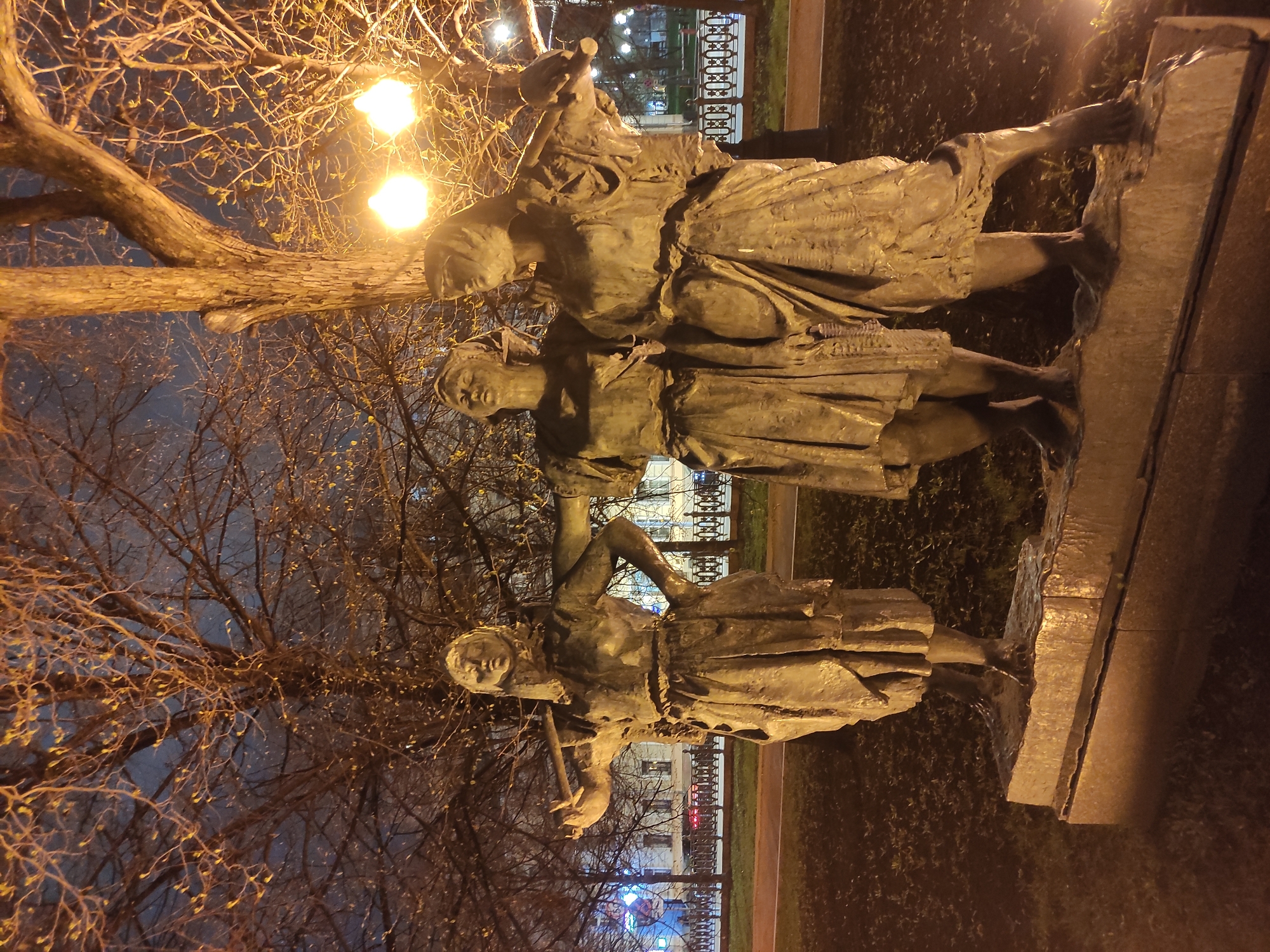 This night-time image, set in Moscow, Russia, displays a monumental bronze statue featuring three female figures. The statue is illuminated by bright, warm-toned streetlights, casting an orange glow on the subjects and the surrounding bare tree branches.
The three figures are depicted standing in a line, dressed in what appears to be traditional or working attire, with drapery suggesting flowing garments. The figure on the right shoulders a bundle, possibly a sheaf of wheat, indicating a theme of harvest or labor. The central figure holds a smaller, indistinct object in her hands, while the leftmost figure holds a long staff or tool, extending downwards. Their expressions are serious and contemplative.
Behind the statue, the intricate, leafless branches of a large tree are visible, with some small buds hinting at early spring. The dark night sky serves as a backdrop. In the mid-ground, a decorative metal fence separates the statue's immediate area from a pathway and further greenery. Distant buildings, softly lit by artificial light, complete the urban setting. The ground around the statue is dark, suggesting grass or low shrubbery. The overall scene conveys a quiet, reflective atmosphere under the artificial glow of city lights. No visible text is present in the image.