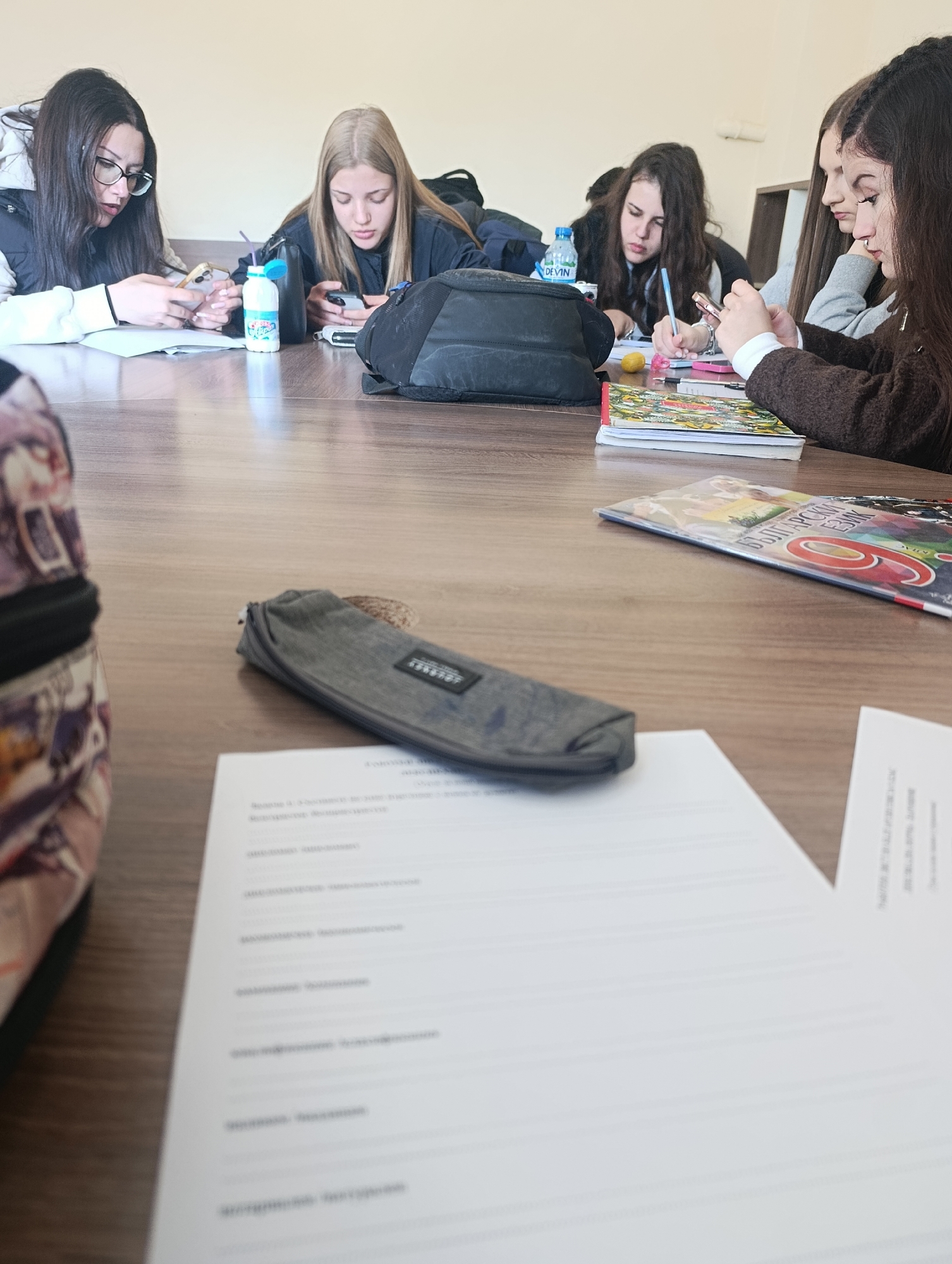 Five young women are seated around a long, light-brown wooden table. They appear to be in a classroom or study hall, possibly in Silistra, Bulgaria, based on the textbook on the table. The women are engaged in various activities: some are looking at their mobile phones, while one is writing. There are several books and notebooks scattered on the table, along with a bottle of water and a pencil case. There are printed papers in front of the table, which are illegible due to the focus of the image. Their attention seems to be divided between their phones and their studies. The lighting appears natural, suggesting the room is lit by daylight.