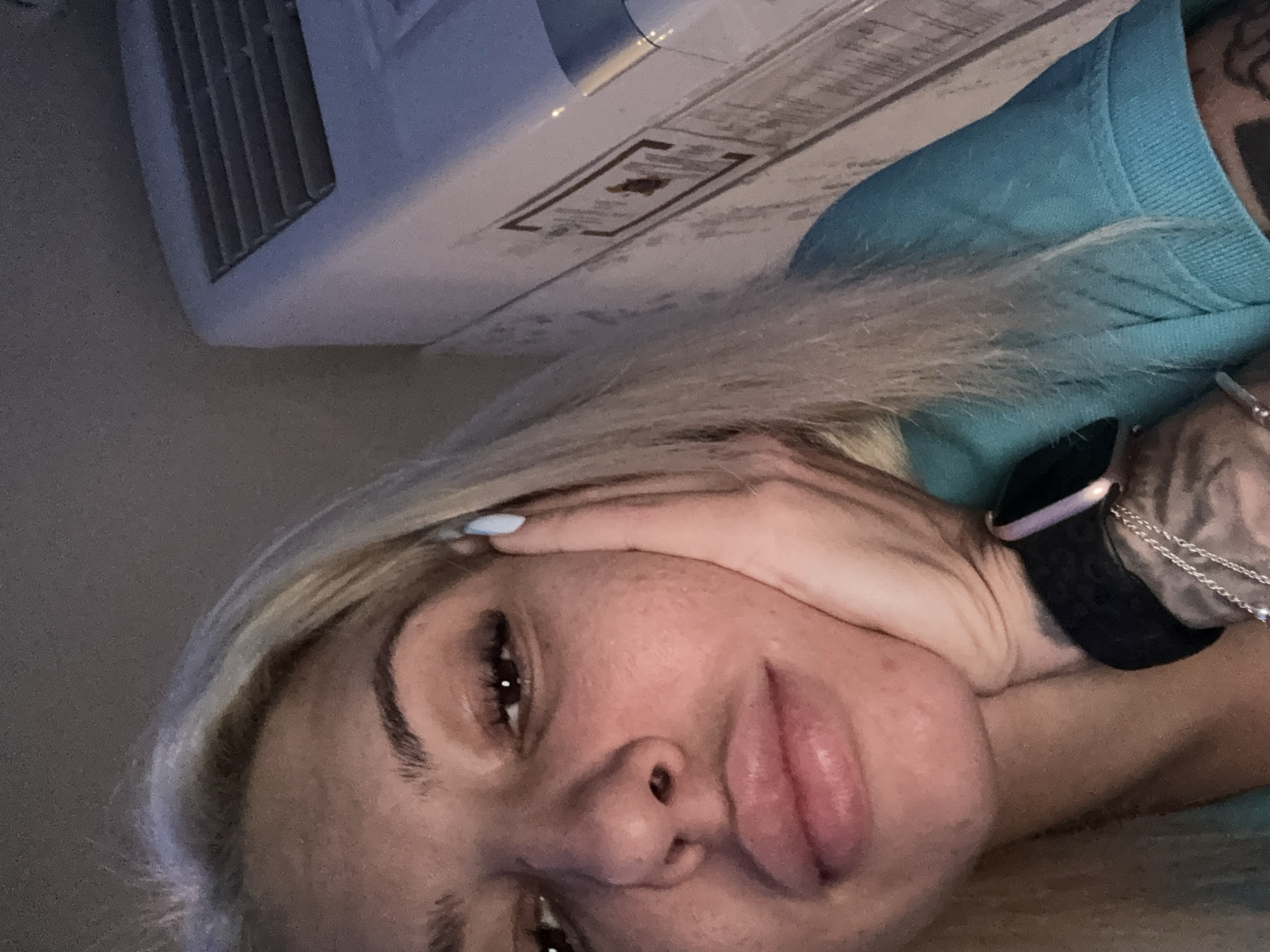 The image shows a close-up, low-angle selfie of a young woman with blonde hair and light skin. She is resting her right cheek on her right hand, which is propped up on her chin and jaw. Her eyes are open, and she is looking slightly to her left. She is wearing a teal-colored shirt and a black smartwatch on her left wrist. Tattoos are visible on her left arm and wrist.
In the background, to the right of the woman, is a white appliance with a gray grille. The appliance appears to be mounted on a wall, and a portion of the wall's textured surface is visible behind it. The lighting suggests an indoor environment, possibly at night or in a dimly lit room. The overall tone of the image is neutral.
Without any identifying landmarks or context, it is impossible to determine the specific location within Plovdiv, Bulgaria, or any other city.