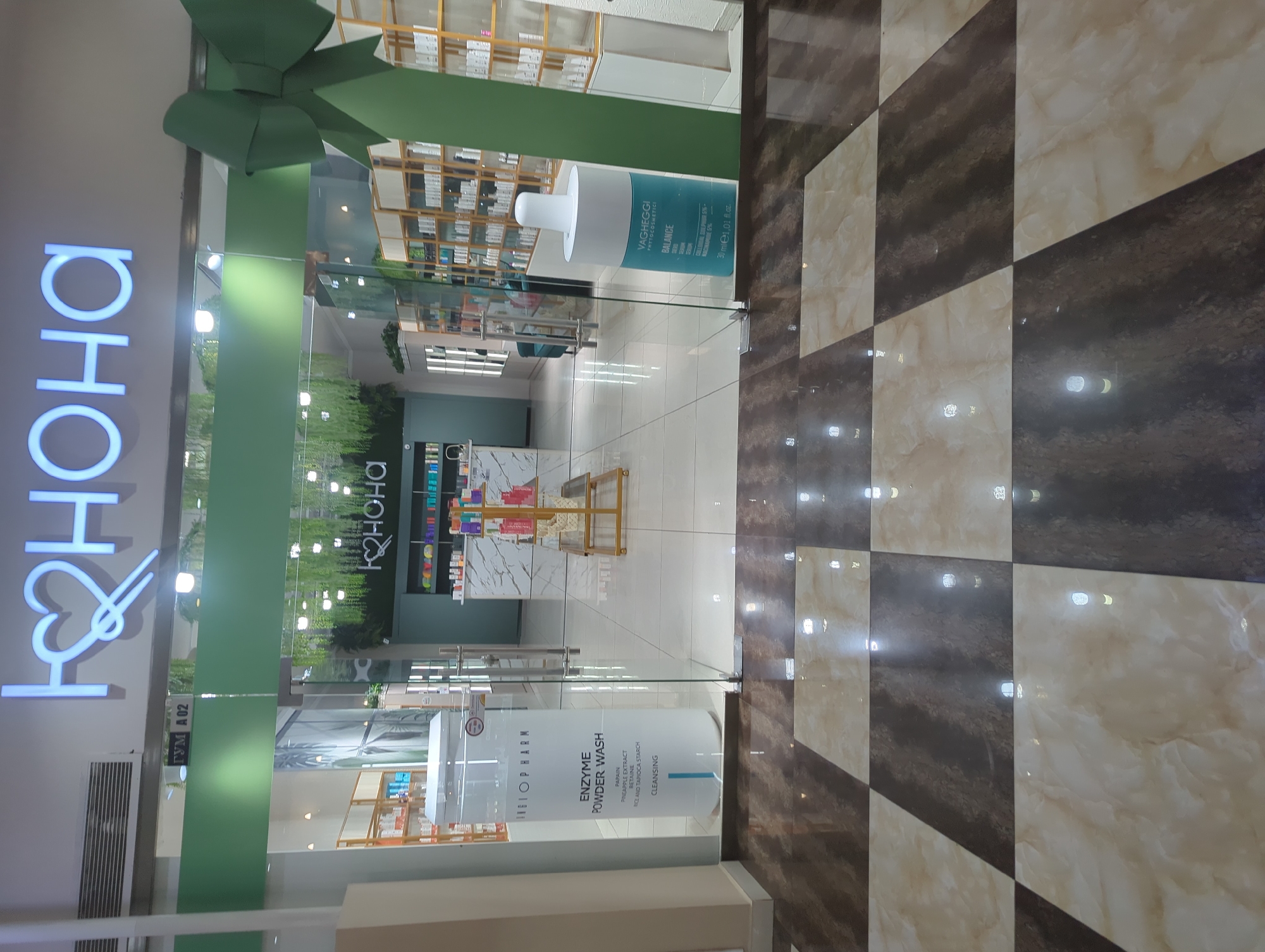 The image depicts the entrance to a store named "HO&HOHA" with a heart symbol in its logo, located in what appears to be a mall or commercial center. The store is decorated with a large green bow. A sign next to the store entrance indicates it is located in the "GYM A 02" area. The interior of the store showcases shelves stocked with various products. Promotional materials, including signs for "INGIOPHARM ENZYME POWDER WASH" and "VAGHEGGI PHYTOCOSMETICI BALANCE" serum, are displayed on the glass entrance. The floor tiles are arranged in a checkered pattern of brown and beige. The location is likely in the city of Bishkek City, Kyrgyzstan.