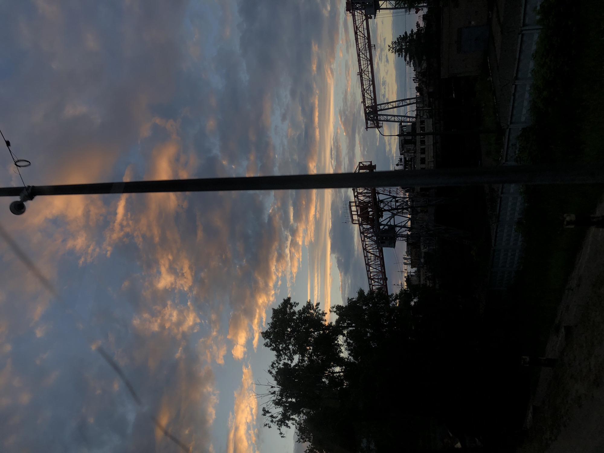A silhouette of trees is visible on the left side of the image, with industrial cranes and buildings in the background. The sky is a mix of blue and orange clouds, indicating either sunrise or sunset. A vertical pole with a light fixture is present in the foreground. The scene appears to be an industrial area, possibly near a port or shipyard, given the presence of cranes and what looks like boats or ships in the background. The lighting suggests it is either early morning or late evening. The location is identified as Chaikovsky, Russia.