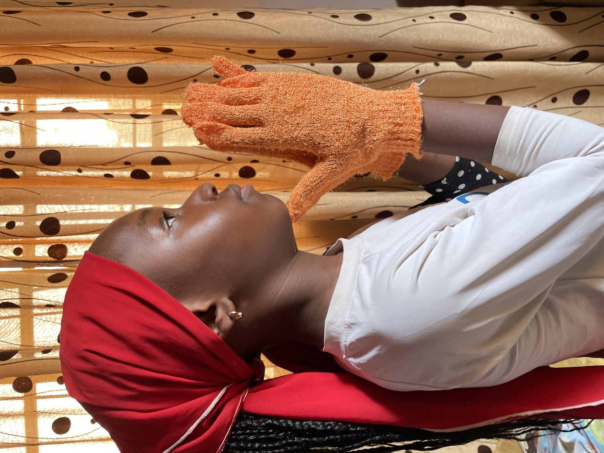 A young woman with dark skin and close-cropped hair is lying down with her head resting on a red cloth. She has a red headscarf wrapped around her head and an earring in her left ear. She is wearing a white shirt. Above her, an orange exfoliating glove is positioned over her face. The background consists of a window with patterned brown curtains. The image appears to be taken indoors, but the location in Jos, Nigeria is not evident from the image itself.