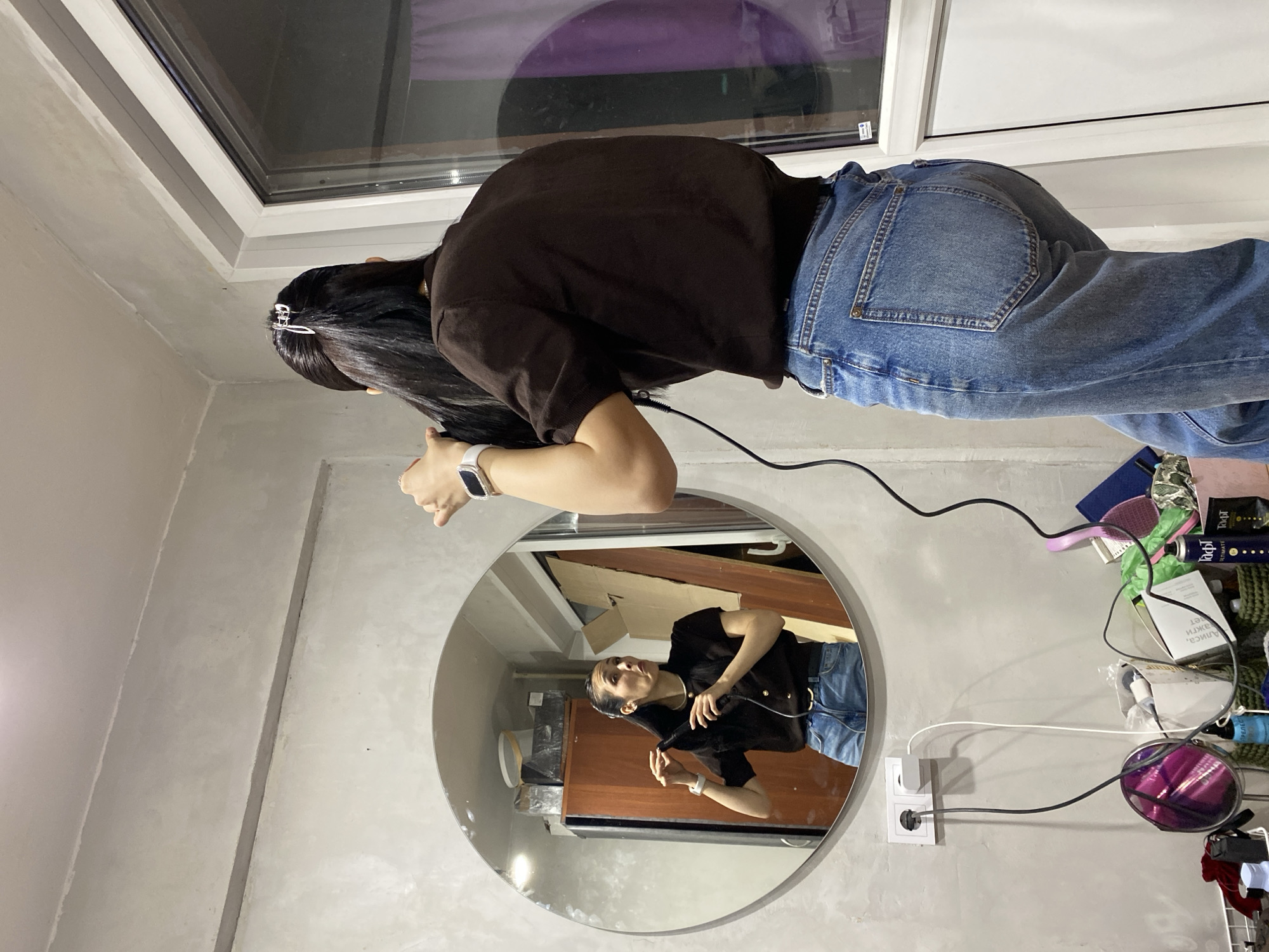 The image, rotated 90 degrees to the left, captures an indoor scene, likely a room in Votkinsk, Russia. A person, viewed from behind and slightly turned, is the main subject. She has long, dark hair styled with a silver hair clip and wears a dark brown short-sleeved top and blue jeans. A white smartwatch is visible on her left wrist, and a black cable extends from her side towards the bottom right of the frame.
The room features light grey, textured walls, possibly concrete or stucco. A window in the upper left shows a dark exterior, indicating it is evening or night. A large, circular mirror is mounted on the wall below the window.
The mirror reflects the same person from a front-facing perspective. In the reflection, she appears to be wearing a black top with distinctive gold buttons and blue jeans. She looks upwards, seemingly engaged in styling her hair or adjusting something near her head. The mirror also reveals parts of the room's interior, including cardboard boxes, wooden furniture (such as a cabinet or dresser), and a white container.
To the right of the mirror, a white double electrical socket is visible, with both black and white cables plugged in. Adjacent to the socket is a cluttered shelf or surface holding various personal items, including a pink hairbrush, bottles (one clearly labeled "Taft"), and other small objects. Reversed text on a white box within the mirror's reflection reads "ЭКСЛЮЗИВ КЛИПСА" (Exclusive Clip), suggesting a product related to hair accessories. The overall setting implies a personal space dedicated to grooming or getting ready, with several electronic devices or tools in use.