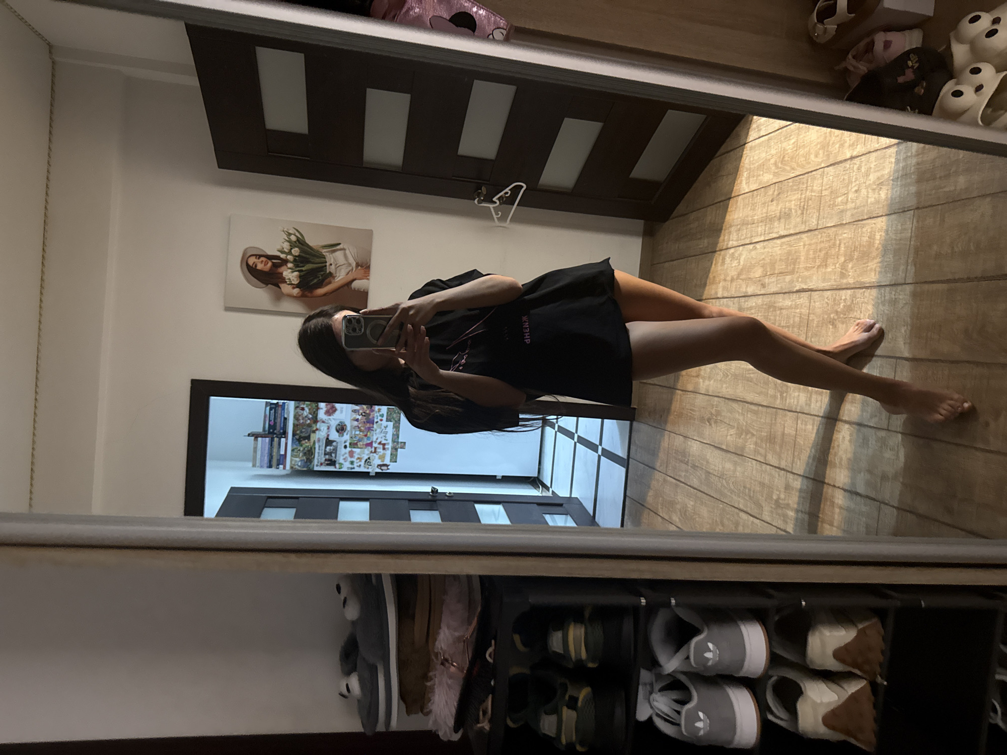 A young woman with long dark hair stands in an apartment and takes a mirror selfie. She is wearing a black t-shirt and white shorts. The woman is holding a smartphone in her right hand and is looking at her reflection in the mirror. In the background, there is a shoe rack with several pairs of shoes, a door, and a painting on the wall. The floor is made of wooden planks. The apartment appears to be located in Chișinău, Moldova.
