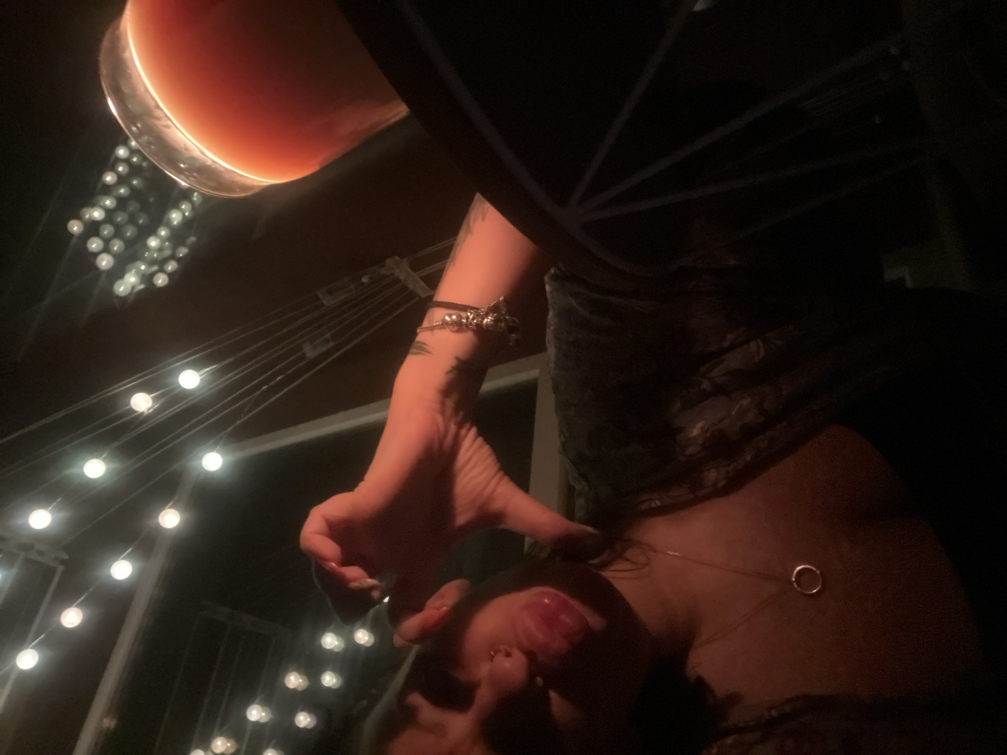 The image shows a close-up of a person's face and hand, forming a heart shape. The person has their lips pursed and is sticking their tongue out slightly. They are wearing a necklace with a circular pendant. In the background, there are strings of lights and a glass of orange liquid. The scene appears to be outdoors at night, possibly at a restaurant or bar. The specific location in the city of Plovdiv, Bulgaria, is not discernible from the image.