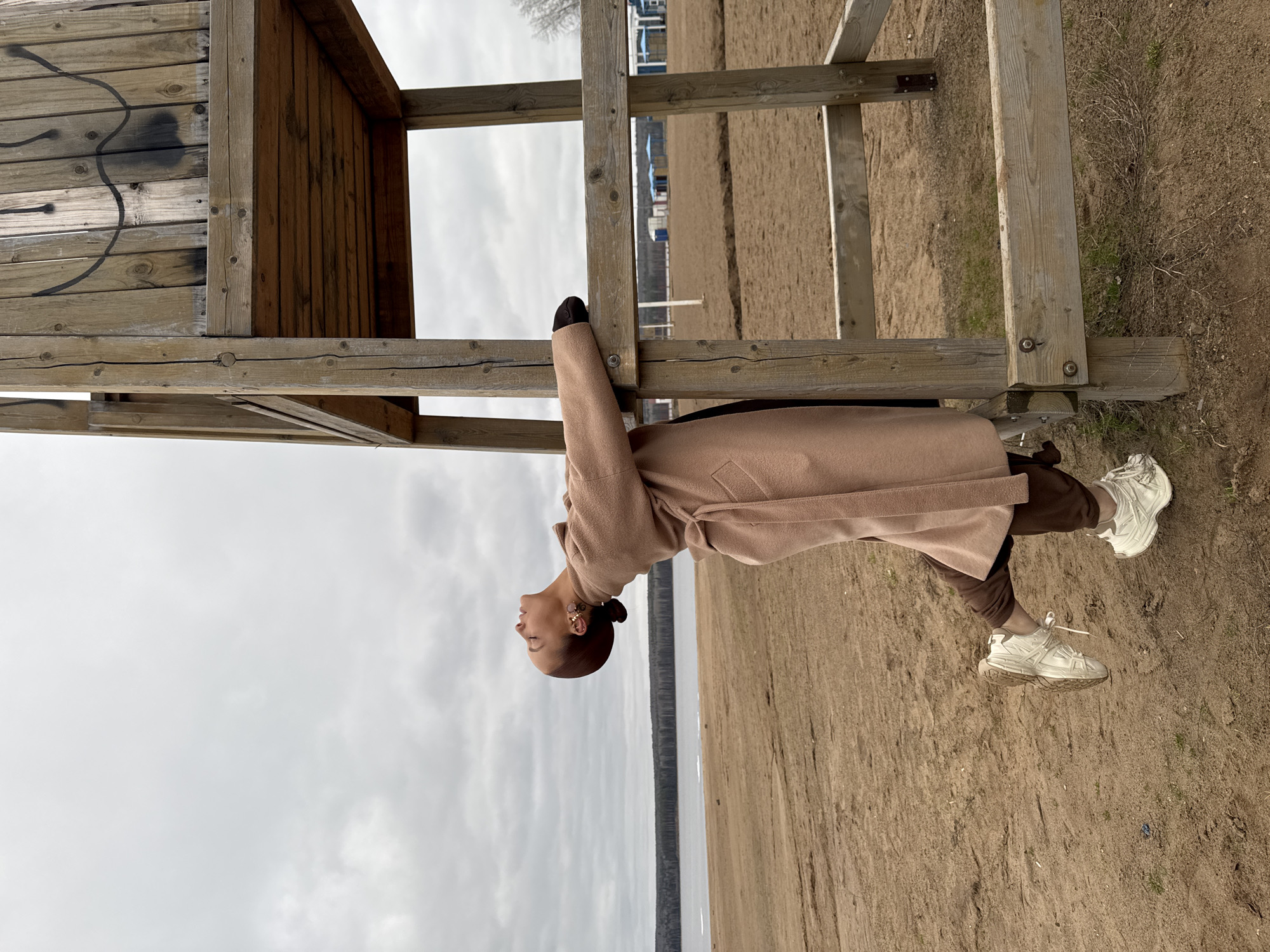 A full-body shot captures a woman posing outdoors, leaning against a large wooden structure. The image is oriented sideways, but if viewed upright, the woman stands vertically, looking upwards with her eyes closed, displaying a serene expression. She is dressed in a long, light beige or camel-colored wool coat, dark trousers, and white chunky sneakers. Distinctive pink and purple earrings are visible.
The wooden structure, possibly a lifeguard tower or observation deck, dominates the left side of the frame. It features horizontal and vertical wooden planks and beams, with some dark, abstract graffiti on one panel.
The setting appears to be a wide, sandy area, likely a beach or riverbank, with sparse dry vegetation. In the background, a large body of water, possibly the Kama River or a reservoir near Chaikovsky, Russia, stretches across the horizon, bordered by a distant treeline and faint structures. The sky is overcast, suggesting cloudy weather conditions during daytime. The overall mood is calm and contemplative. No discernible text is visible.