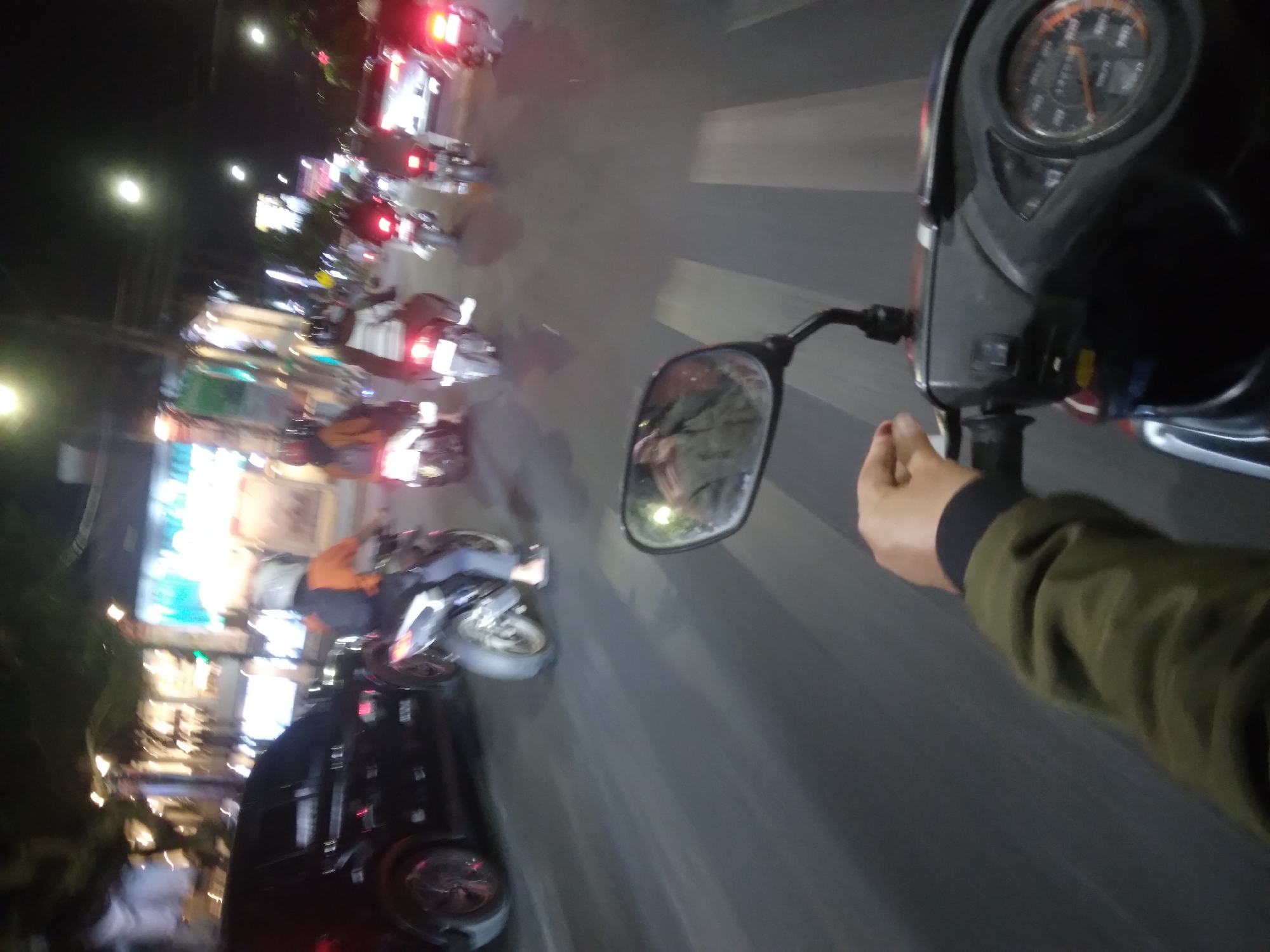 The image is a first-person perspective of a person riding a motorcycle at night through what appears to be a busy street in Purwokerto, Indonesia. The rider's left hand is visible on the handlebars, and their arm is extended forward, wearing a green jacket. The motorcycle's instrument panel is also visible, showing a speedometer. In front of the rider, several other motorcycles are moving in the same direction, some carrying passengers. The motorcycles have their brake lights illuminated, indicating traffic. To the left, a black car is visible. The road has pedestrian crossing markings. The background shows buildings with illuminated signs and streetlights, suggesting an urban commercial area. The overall scene depicts nighttime traffic congestion.