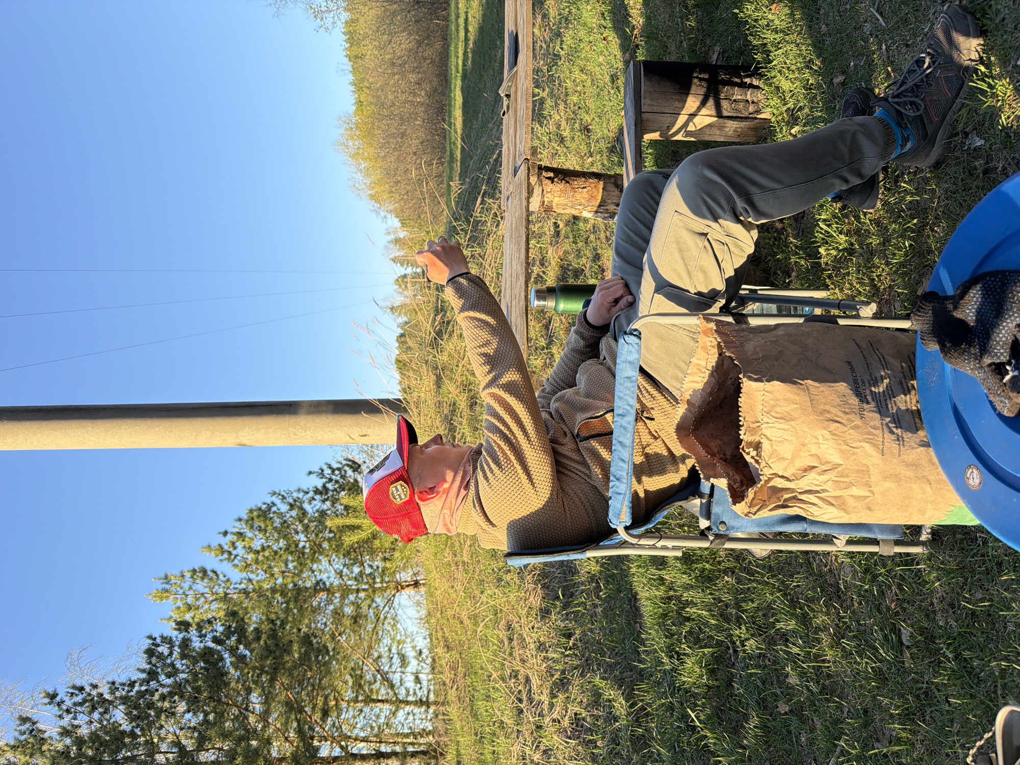 A person in a red and white hat, a patterned jacket, and green pants sits in a blue folding chair outdoors. The person is pointing upwards with their right hand. Beside the chair is a large brown paper bag. In the background are trees, green grass, a wooden fence, a log, and a blue sky. Overhead power lines are visible in the sky. The image location is in the city of Chaykovsky, Russia.