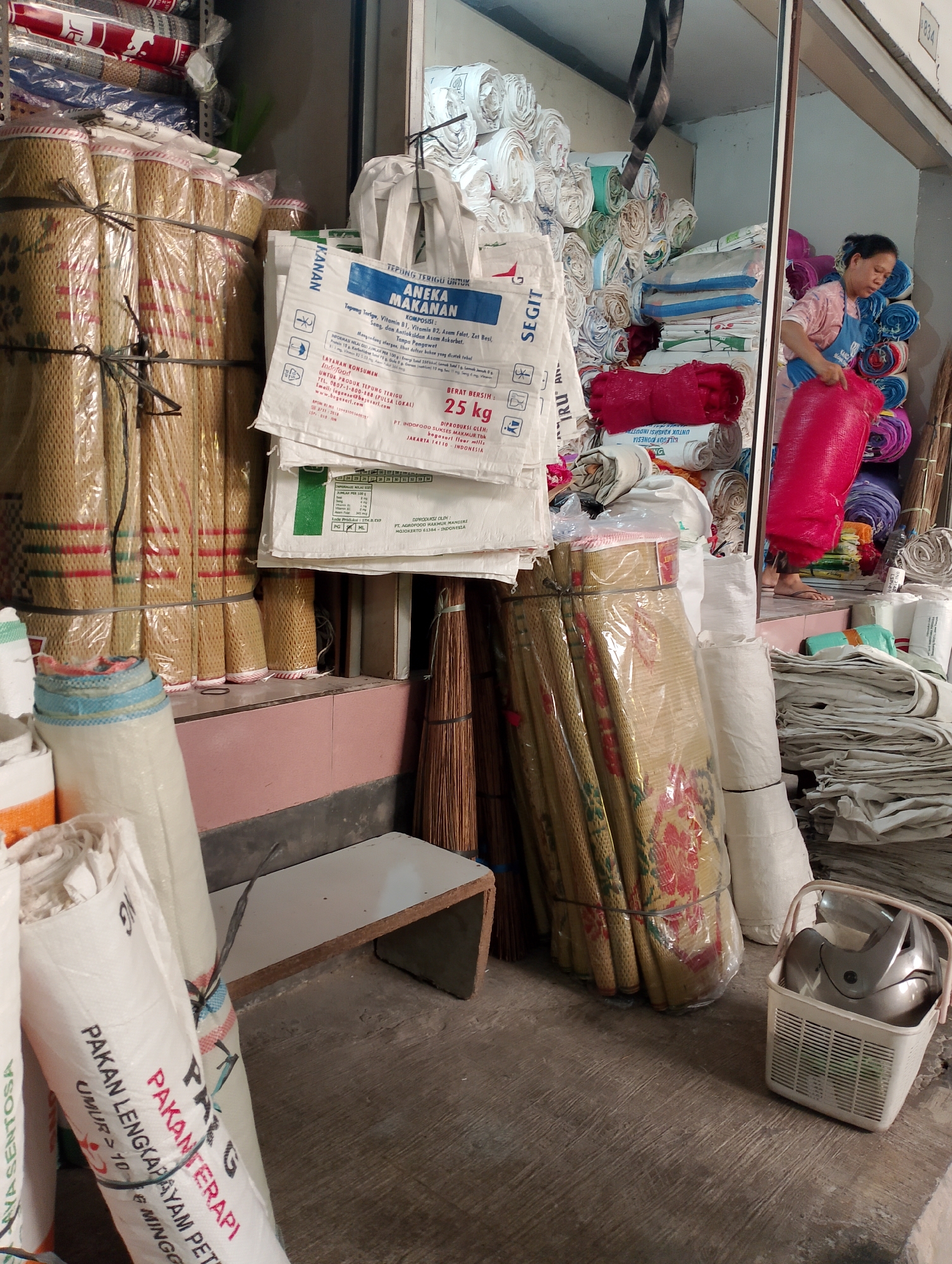 The image depicts an indoor scene in what appears to be a shop or storage area in Magelang, Indonesia, filled with various rolled goods and bags. In the foreground, several large rolls of woven mats, likely for flooring or seating, are stacked and wrapped in clear plastic. To the left, more rolls of similar materials are visible, some with red and white patterns. White and multicolored sacks, some bearing Indonesian text related to animal feed, are also prominent.
In the mid-ground, a woman wearing a pink shirt and a blue apron is seen in the process of handling a large, bright pink bag. She is standing near a stack of more rolled items, some appearing to be made of fabric in various colors, including purple and blue. To the right of the woman, a white plastic basket containing a silver object, possibly a helmet or a car part, is placed on the floor. Further to the right, a pile of folded white fabric or tarpaulin is visible.
The environment suggests a commercial or industrial setting. The floor is concrete, and the walls are a neutral color, with some exposed metal structures in the background. The lighting appears to be artificial, with some natural light possibly entering from an open doorway or window on the right side of the image. The overall impression is one of a busy storage or sales area for household goods or materials.