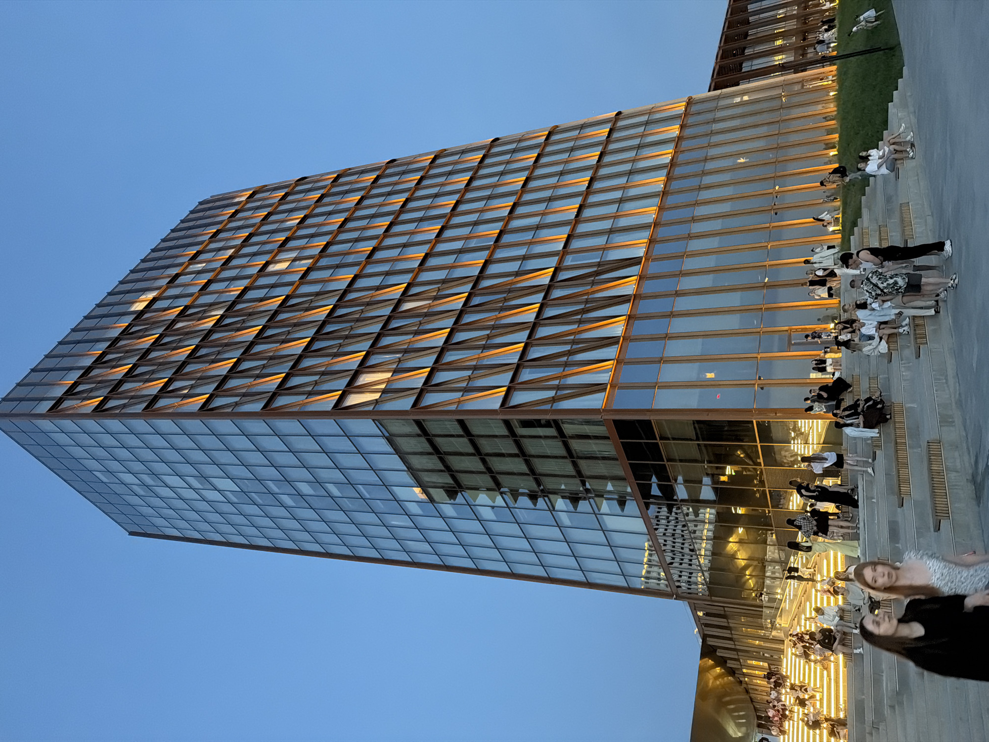 The image depicts a modern building with a glass facade, located in an urban environment. The building appears to be a high-rise structure with a distinct grid-like pattern formed by its windows. A group of people are gathered on a stepped area or plaza adjacent to the building. The sky is clear and blue, suggesting either early morning or late afternoon. The building's facade reflects the surrounding environment. The stepped area where people are gathered appears to be illuminated with soft, warm lighting.