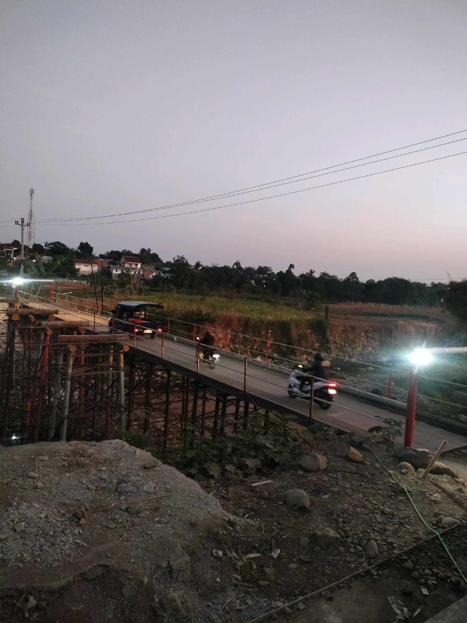 The image depicts a bridge construction site near a rural area, likely at dusk or dawn. A partially constructed bridge is visible in the foreground, with scaffolding and materials indicating ongoing work. A paved road spans the bridge, with vehicles and a motorcycle in motion. A car is driving across the bridge, and a motorcycle is following behind. The surrounding environment includes vegetation, some housing in the distance, and power lines. The sky is a pale gray, suggesting low light conditions. The overall scene is one of activity and development in a natural setting. The location is identified as Tersono, Indonesia.