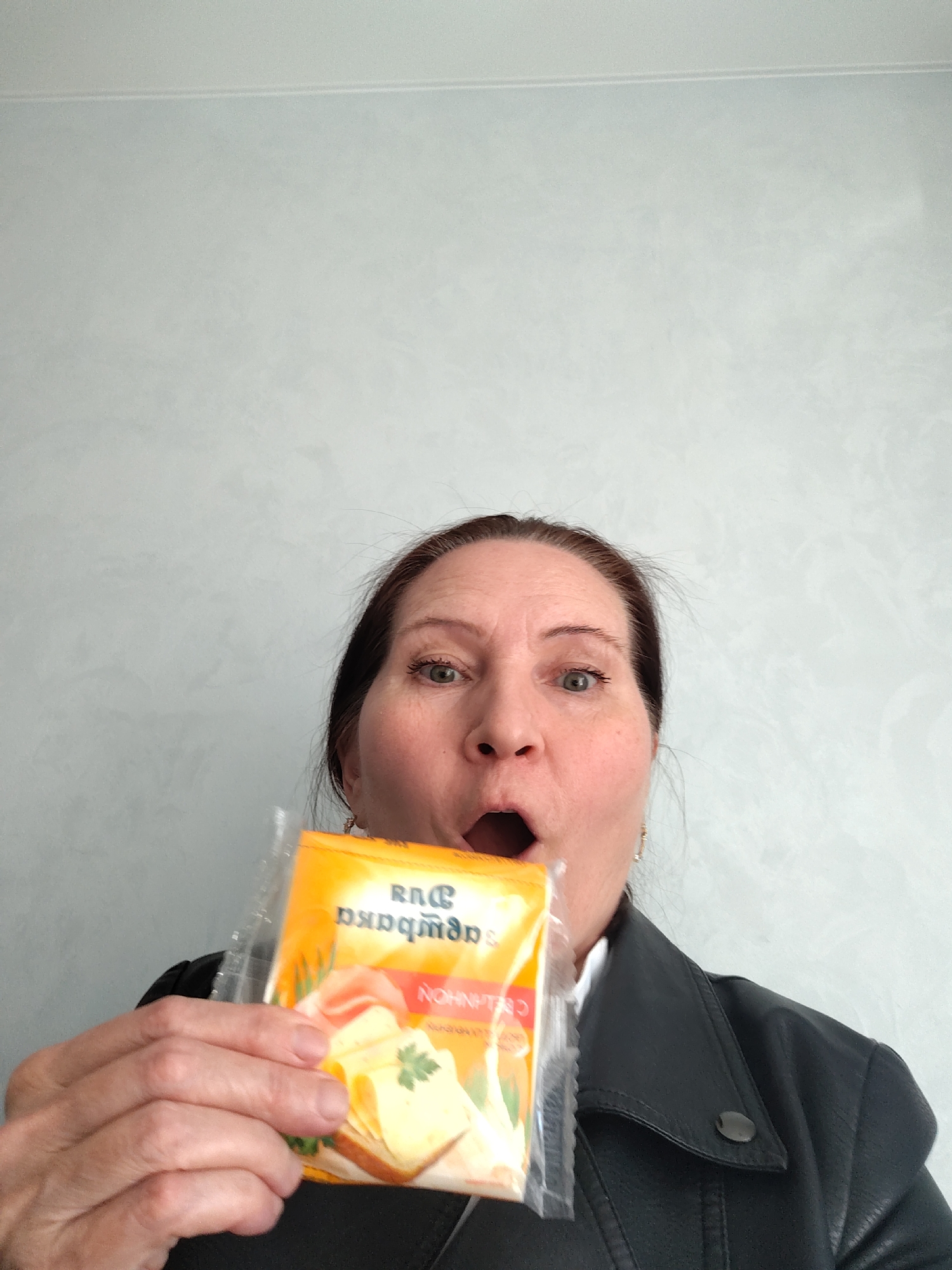 A woman is holding a package of processed cheese in front of her face. She has an open mouth expression. The package is yellow and contains a picture of a cheese sandwich. The woman is wearing a black jacket. The background is a light gray wall. The city is not visibly identifiable in the image.