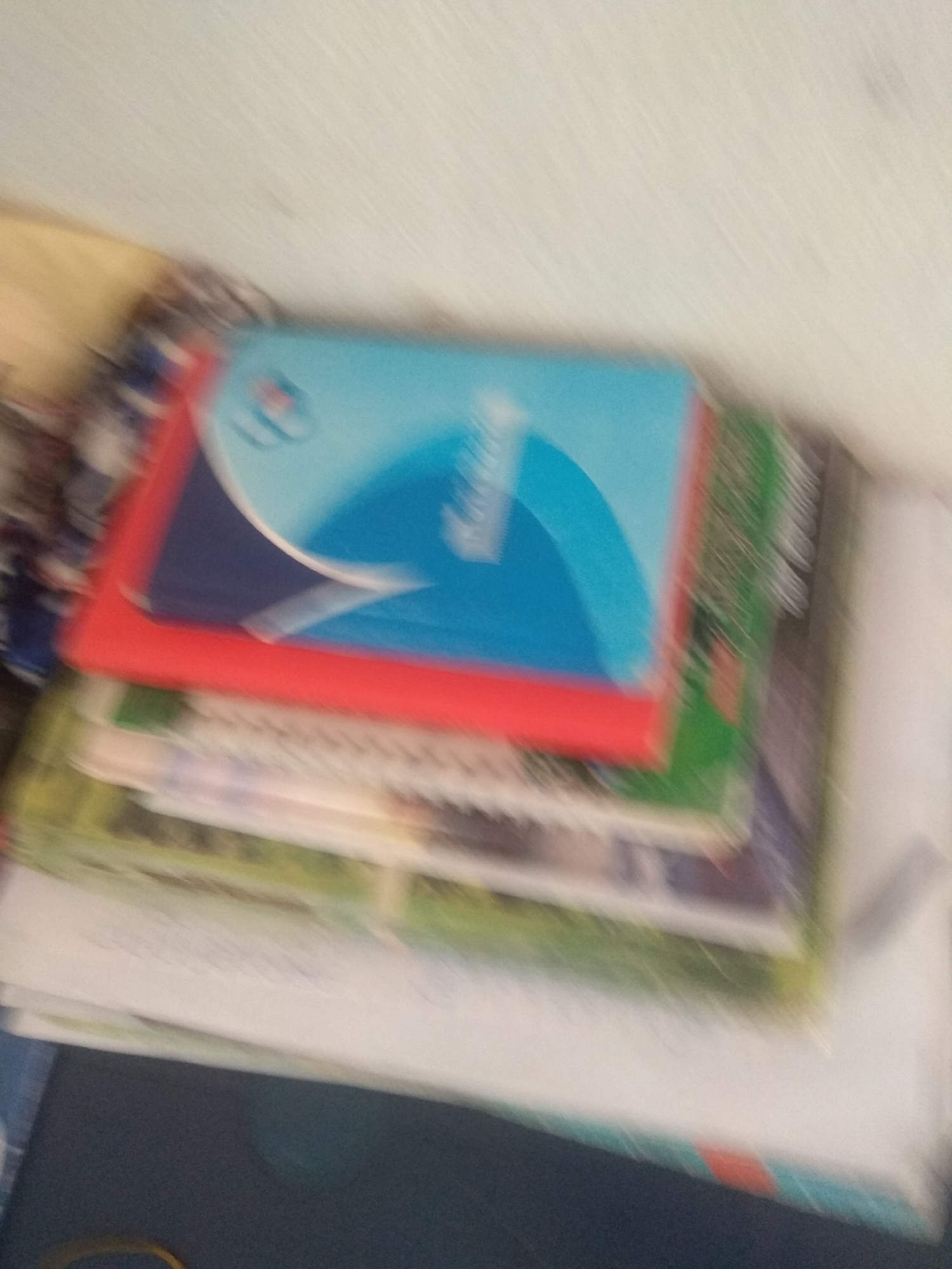 The image shows a stack of books and papers, mostly out of focus. The topmost item is a book or notebook with a blue and white cover, featuring a logo that appears to be a stylized "P" inside a circle. It has a red border. Beneath this are several other books or papers with visible green, white, and possibly other colors. The overall setting appears to be indoors, likely on a surface like a table or shelf, with a light-colored wall in the background. The image quality is poor due to blur and low resolution, making it difficult to discern specific details or identify any people. There is no visible indication of the specific location within Jalingo, Nigeria.