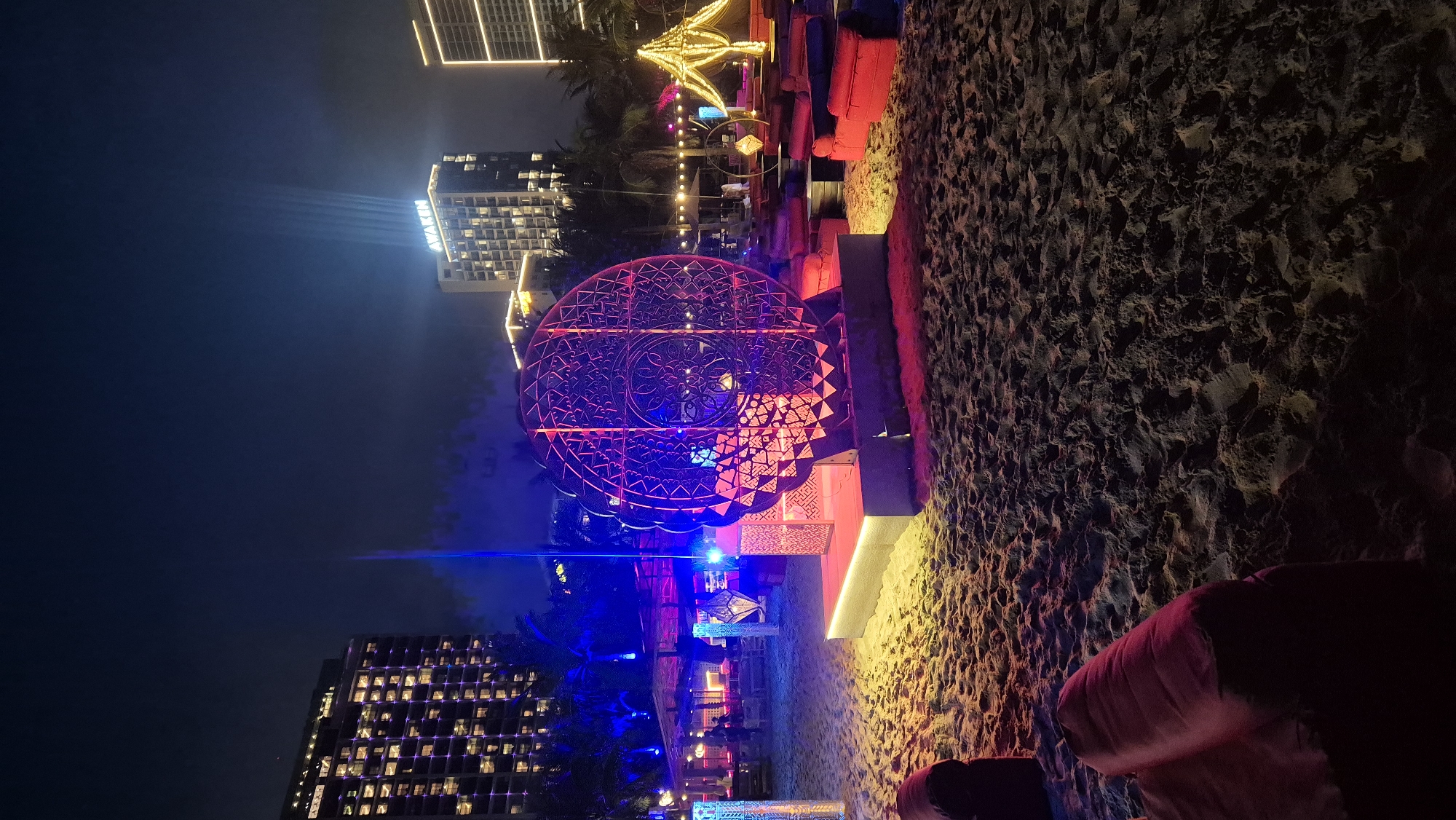 The image captures an outdoor nighttime scene on a sandy beach, likely at a resort or event space in Da Nang, Vietnam. In the foreground, there are plush, red seating cushions scattered on the sand. The midground features a large, circular metal art installation with intricate geometric patterns, illuminated by red and purple lights. To the left of this sculpture, there are more decorative elements, including illuminated pillars and possibly a bar or seating area with people faintly visible. In the background, modern high-rise buildings are lit up, with one building displaying the word "AWAKEN" in prominent signage. The sky is dark, and beams of light are projected upwards from the buildings. String lights are also visible, hung over seating areas and a draped tent-like structure. The overall atmosphere is festive and illuminated, suggesting a night-time event or entertainment venue.