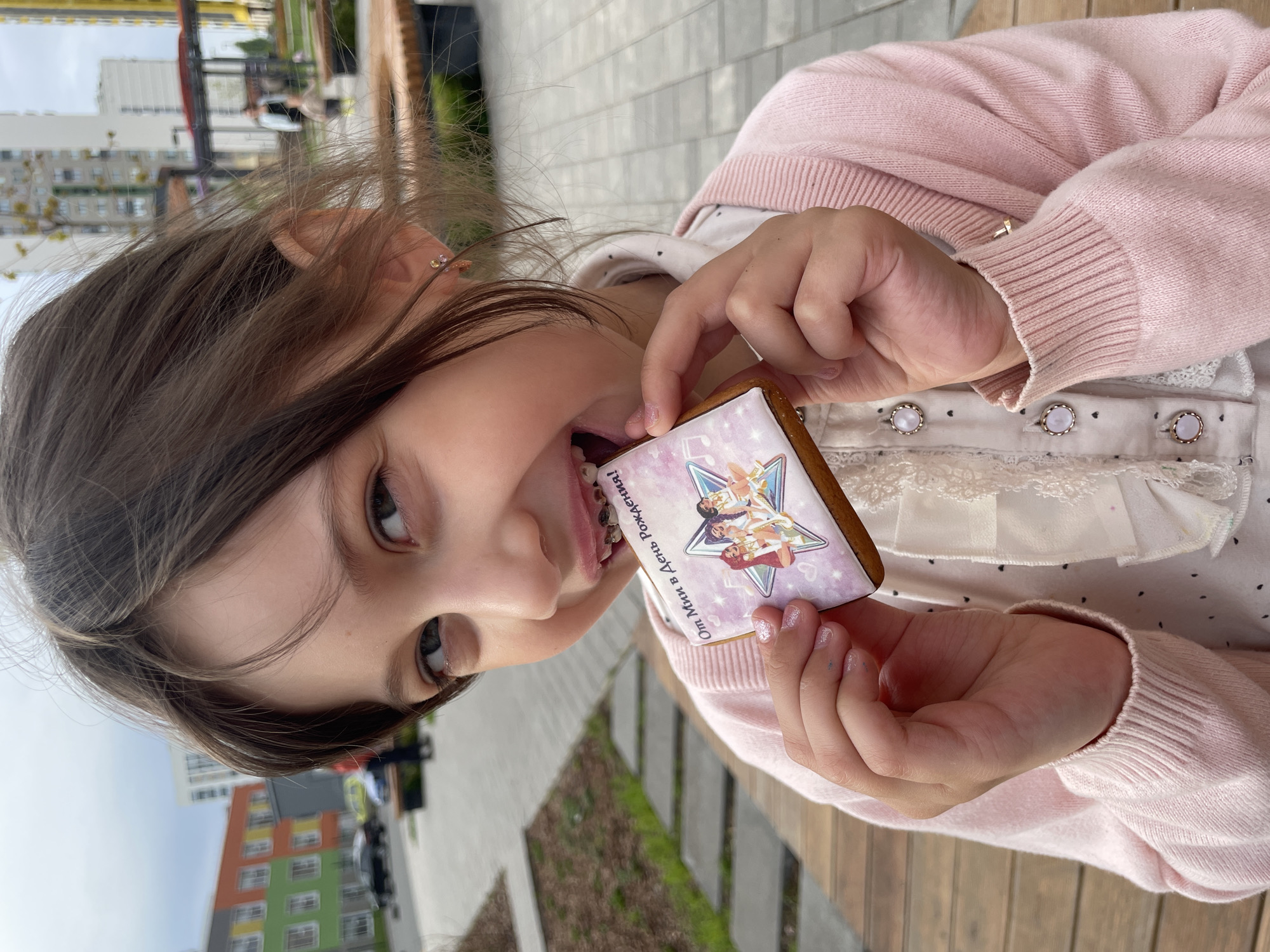 A young girl is biting into a square gingerbread cookie with an image printed on it, held in her hands. She has brown hair and is wearing a pink cardigan over a white, lace-trimmed shirt. Her teeth are visible. The background shows a paved area with wooden seating and some greenery. In the distance, there are buildings, including a colorful apartment building. The city is Izhevsk, Russia.