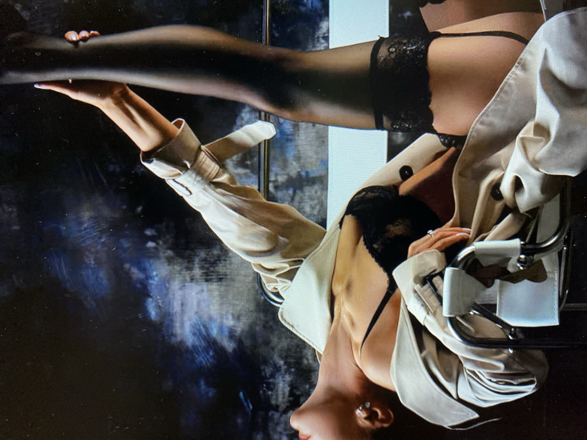 A person is lying on their back, with their head and torso tilted back and to the left. Their right leg is extended upwards and to the right, bent at the knee. They are wearing a beige trench coat, a black lace bra, and black stockings with lace trim. Their left arm is bent and raised, with their hand touching their extended right leg. Their right arm is bent and resting on their chest. They are wearing a ring on their right ring finger. The background is dark and textured, with shades of blue and grey. The person is lying on a chair with chrome legs and white leather accents. The lighting is dramatic, casting shadows and highlighting certain areas of the body.