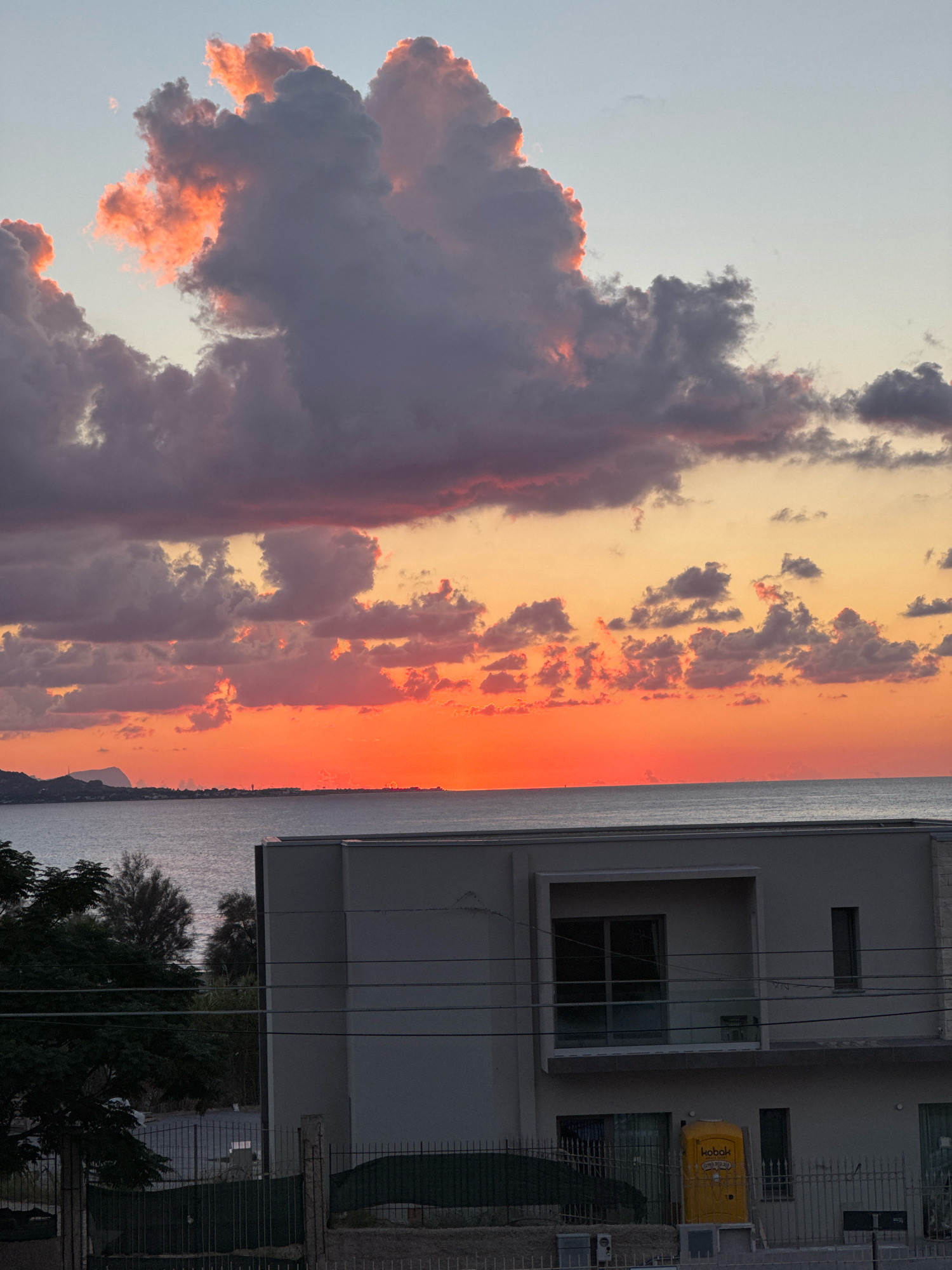 The image depicts a sunset over the sea in Capaci, Italy. The sky is filled with dramatic clouds, illuminated by the setting sun in vibrant shades of orange and pink. The sea is calm, with a distant coastline visible on the horizon, featuring hills and some structures. In the foreground, a modern building with a balcony and large windows is partially visible. A yellow portable toilet stands near the building. Trees and a fence are also present in the foreground, suggesting a residential or construction area. The overall scene conveys a serene and picturesque view of the Italian coast at dusk.