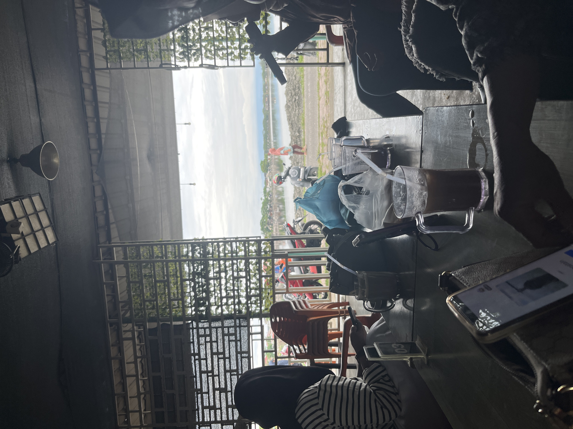 The photograph depicts a scene at a roadside cafe in Lhokseumawe, Indonesia. The view overlooks a coastal area with a cloudy sky. Several motorcycles are parked near the waterfront.
In the foreground, people are sitting at tables. A glass of iced coffee and a smartphone are visible on one table. Another person is wearing a striped shirt and appears to be looking at a phone. A stack of orange plastic chairs is placed nearby. The scene is partially obscured by a metal lattice structure, common in Indonesian roadside cafes.