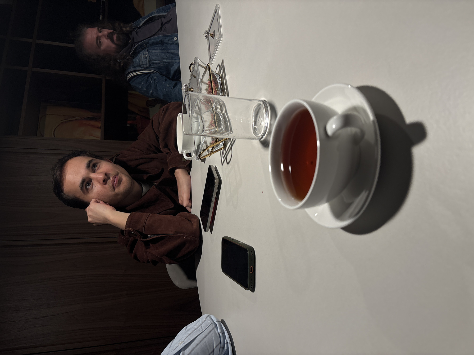 Two men are seated at a white table in an indoor setting, possibly a cafe or restaurant. One man, wearing a brown jacket, rests his head on his hand. A cup of tea rests on a saucer beside him. A clear glass sits on a coaster beside the cup. Two cell phones also sit on the table. The second man, visible in the background, has a beard and wears a blue denim jacket. The location may be Moscow, Russia.