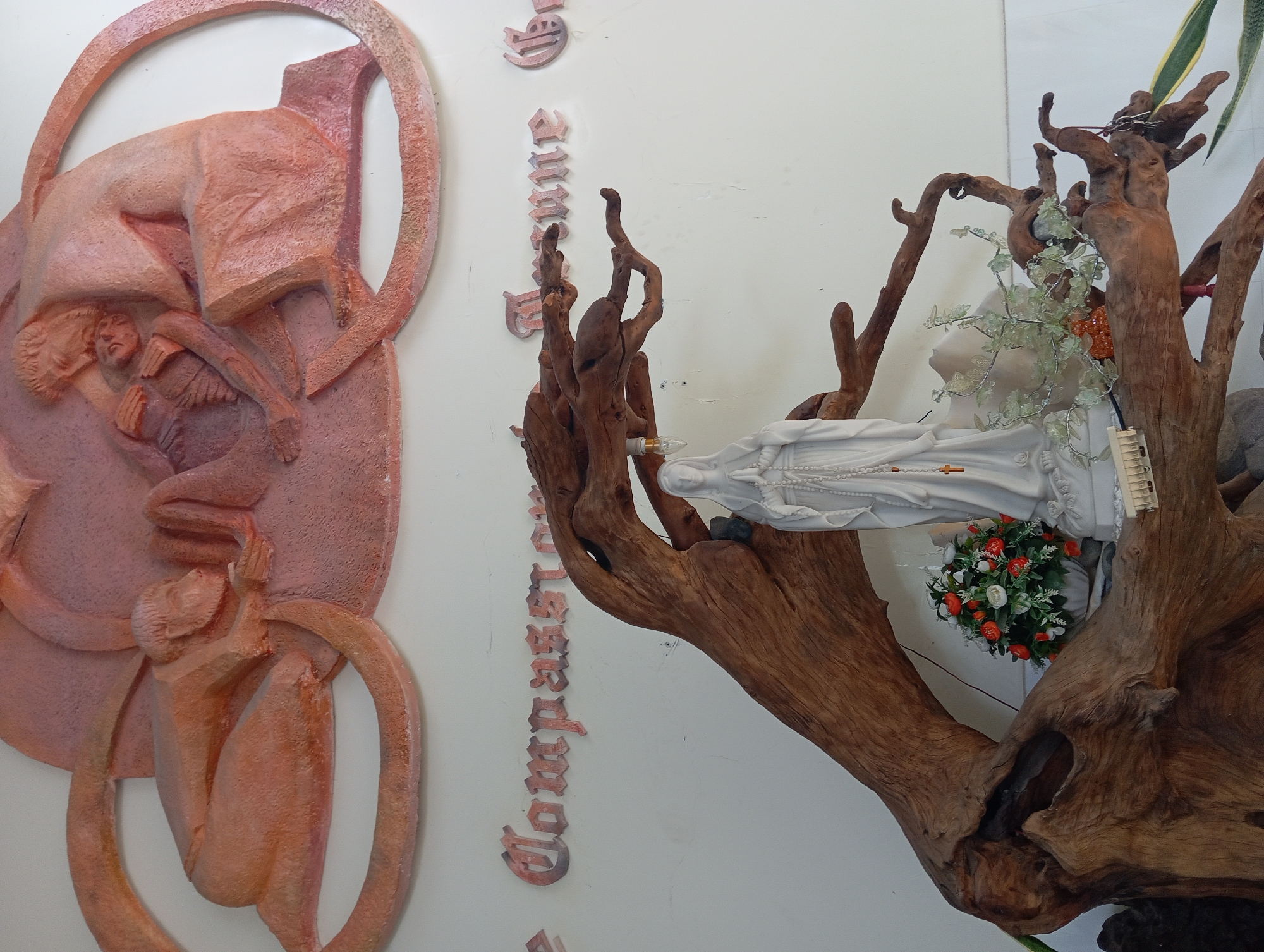 This is a photograph taken in Nha Trang, Vietnam, of a religious display. In the upper part of the image, there is a relief sculpture depicting figures, likely religious in nature, rendered in a pinkish-orange material. Below this sculpture, on a white wall, are words in stylized lettering; partially visible are "Compassion" and "Mercy".
The focal point of the lower half of the image is a statue of the Virgin Mary, made of white material, standing amidst an arrangement of dried wood. The wood forms an organic, branching structure that frames the statue. To the left of the statue, a small vase holds a bouquet of artificial flowers, predominantly red and white. To the right of the statue, some decorative foliage and small orange crystals are arranged on the wooden structure. A lightbulb hangs from one of the wooden branches. The overall scene appears to be an indoor religious installation.
