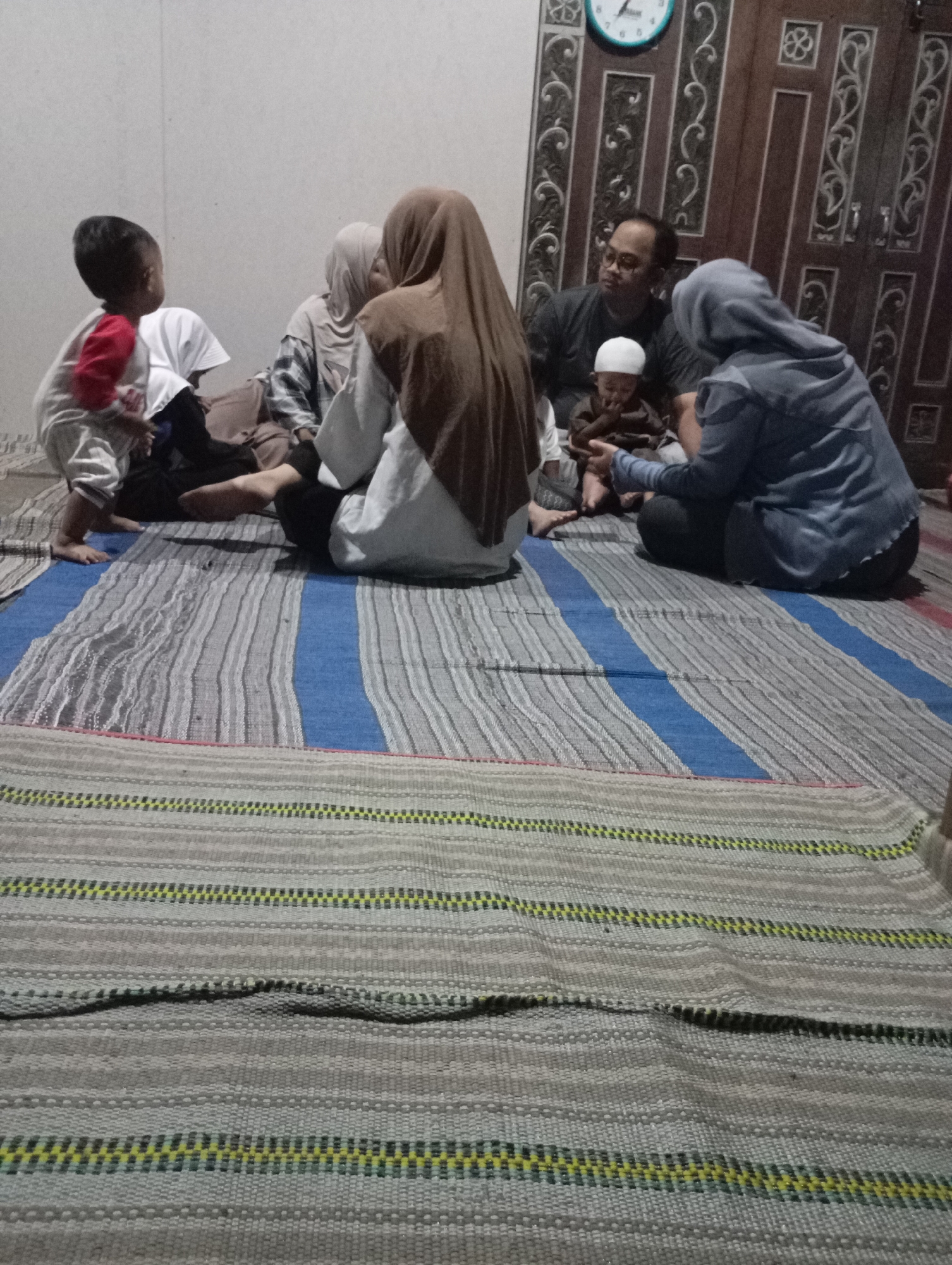 In Ketepung, Indonesia, a group of people are seated on a striped woven mat. In the foreground, the mat is a blend of grey and white horizontal stripes, with a thicker band of blue and white stripes further back. A young child wearing a red and white shirt is seated on the left, facing away from the camera. Beside them are two adults, one wearing a light-colored headscarf and the other a brown hijab. Further to the right, an adult male with glasses is seated and looking towards a baby in a dark outfit and white cap, who is being held by an adult wearing a blue hijab. In the background, a wooden door with ornate carvings and a clock are visible. The lighting suggests an indoor setting, possibly a living room.
