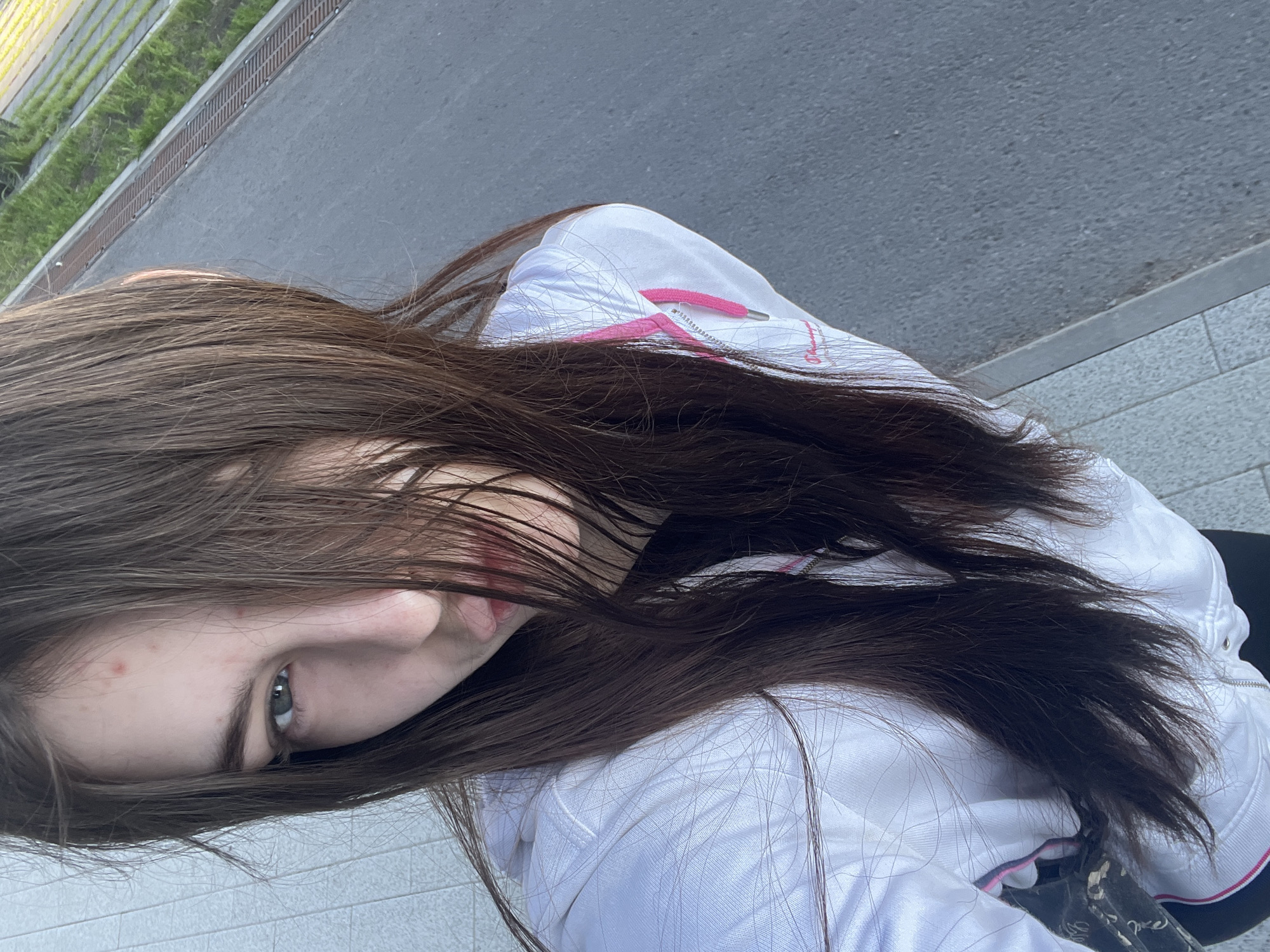 The image captures a close-up, tilted perspective of a young person, likely female, with long, dark brown hair cascading over her face. Her left eye (from the viewer's perspective) is visible and appears green or hazel, looking directly towards the camera. Part of her nose, cheek, and lips are also visible, with a couple of small reddish blemishes on her cheek.
She is wearing a white long-sleeved top, possibly a jacket or hoodie, which features pink details such as a drawstring and a stripe or lining near the collar/zipper area. A dark, possibly black, garment is visible underneath her white top.
The setting is outdoors during the daytime. The background primarily consists of a grey asphalt road, bordered by a dark grey curb and a lighter grey tiled or paved surface, indicative of a sidewalk or pedestrian area. A metal grate or drainage system runs alongside the road, next to some light green vegetation, possibly grass or bushes. The lighting appears soft and diffused, suggesting an overcast day or indirect sunlight. The unusual angle of the photograph gives the impression that the subject is looking up while the camera is positioned above, or the image itself is significantly rotated. No text is visible in the image.