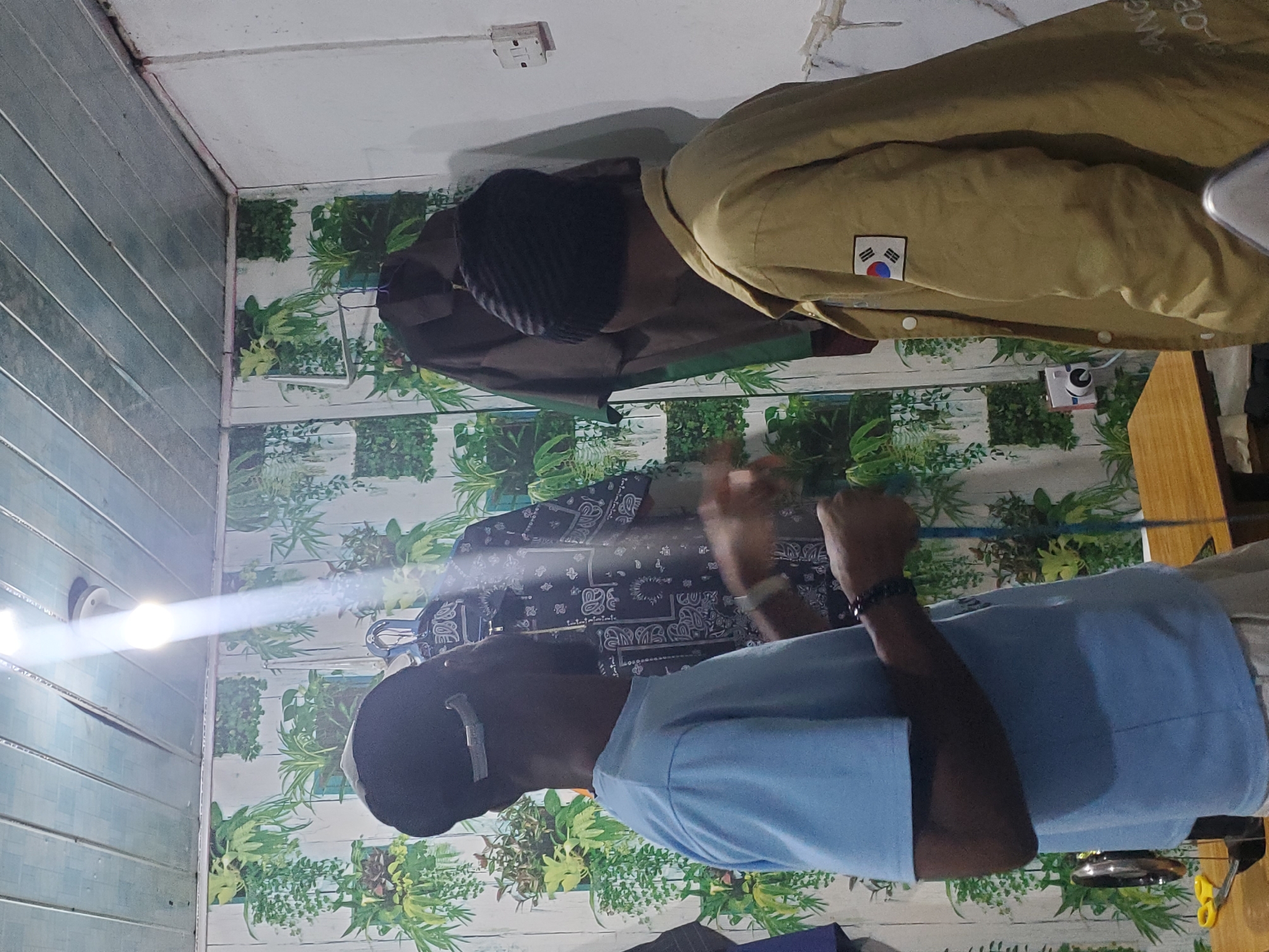 Two young men are standing in a room. The man on the left is wearing a light blue t-shirt and a black cap. He is holding a measuring tape in his right hand and appears to be explaining something to the man on the right, who is wearing a black beanie and a tan jacket with a South Korean flag patch. The room has patterned wallpaper with green plants and a wooden ceiling. There is a sewing machine and scissors on a table. This scene likely depicts a tailor's workshop or a clothing design studio in the city of Jos, Nigeria.