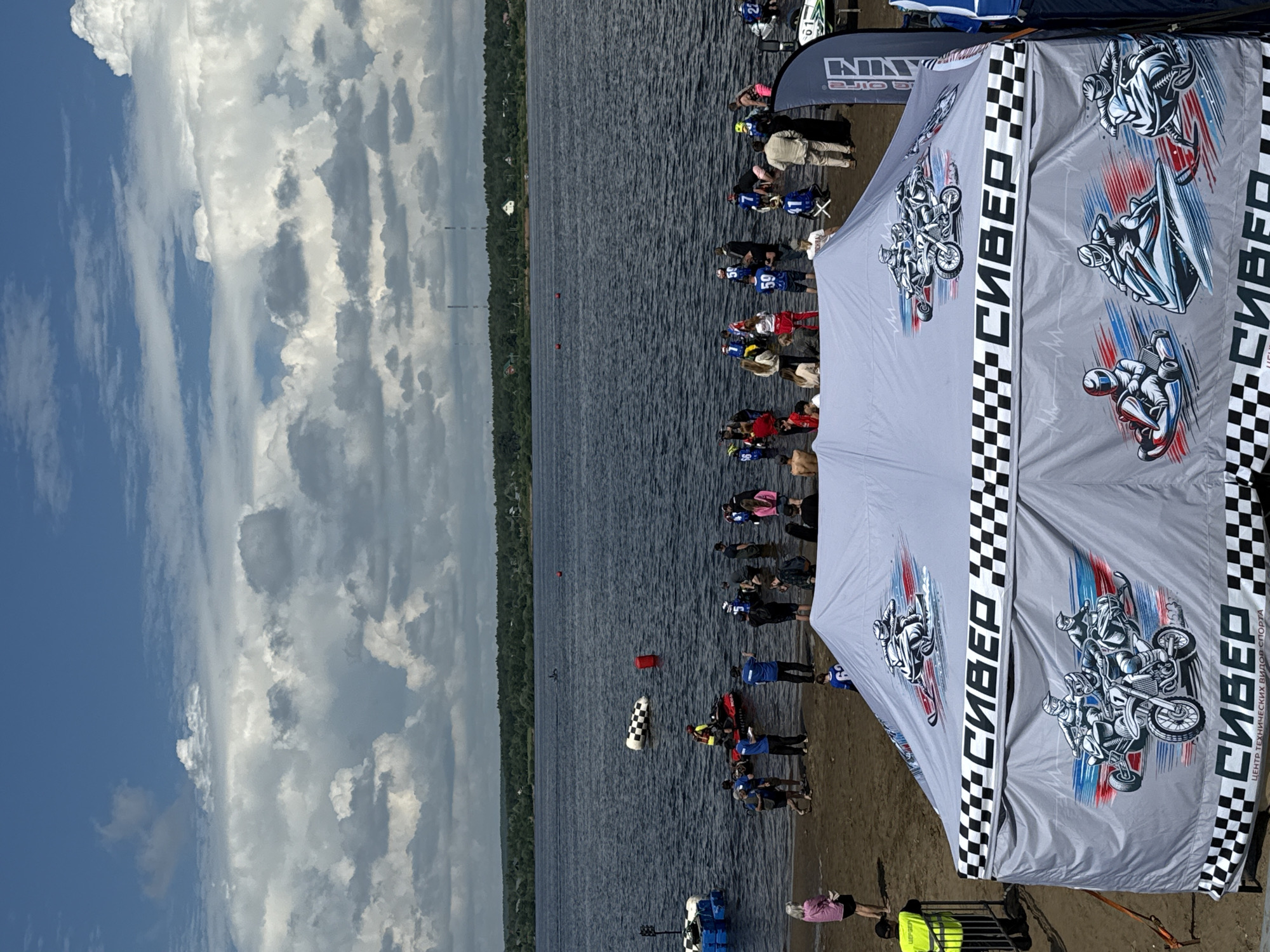 An outdoor scene features a body of water, likely a river or lake, under a cloudy sky with patches of blue. The foreground is dominated by a large, light-colored tent with checkered trim, displaying graphic illustrations of various racing vehicles, including motorcycles, jet skis, and snowmobiles, along with Russian text. A sandy shoreline extends from the tent towards the water, where numerous people are gathered, many wading in the shallows.
In the water, several individuals are on or around jet skis, and a person in a yellow vest is positioned on one. A checkered buoy and a red marker float in the water. Further out in the water, a person is visible on a jet ski. The background shows a tree-lined shoreline with some buildings and distant transmission towers, characteristic of the city of Perm, Russia. The overall atmosphere suggests an event related to water sports or a racing competition.