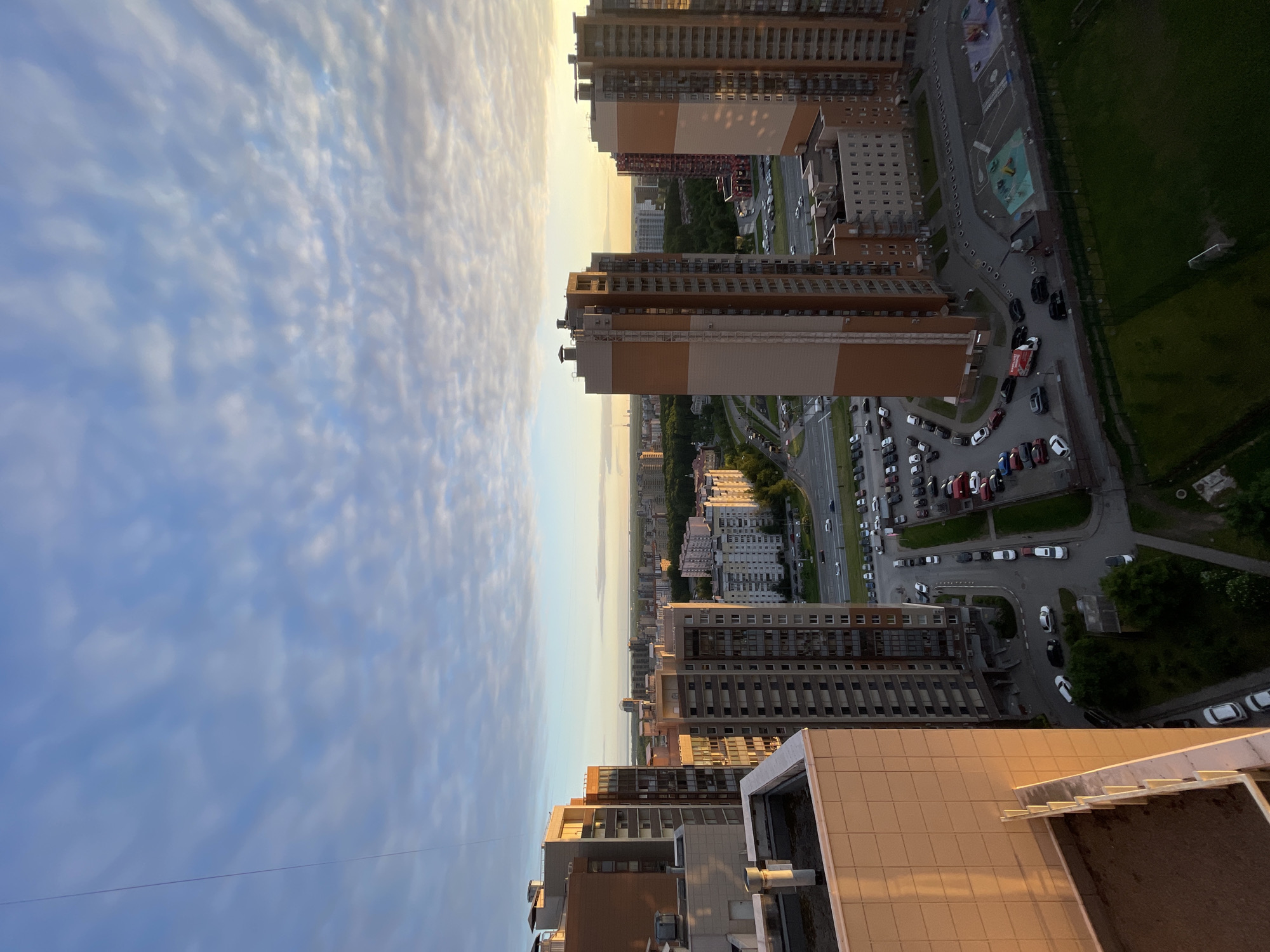 This elevated panoramic shot captures a sprawling modern urban landscape, likely in Saint Petersburg, Russia, during the golden hour or late afternoon. The scene is dominated by numerous high-rise residential buildings and a dynamic sky.
The upper portion of the frame features a vast sky filled with soft, wispy clouds. The clouds near the horizon on the right are bathed in a warm, golden-yellow light, indicative of the low sun, which gradually transitions to a lighter blue on the left. This warm light illuminates the upper sections and facades of the buildings facing the sunset.
The urban environment below consists of multiple multi-story apartment buildings, varying in height and design, with some featuring balconies and distinct brown, beige, and grey paneling. Winding through these structures are roads and streets, bustling with parked and moving vehicles. Numerous cars fill parking lots and line the roads, suggesting a densely populated area. A red truck with indistinct branding is visible near one of the parking areas.
Interspersed among the buildings and infrastructure are green spaces, including lawns and trees. A clear football field with a visible goalpost can be seen on the right side of the image, adjacent to a colorful children's playground. The overall impression is a vibrant, contemporary city scene under the soft, warm light of a setting or rising sun. No discernible text is visible on any signs or vehicles.