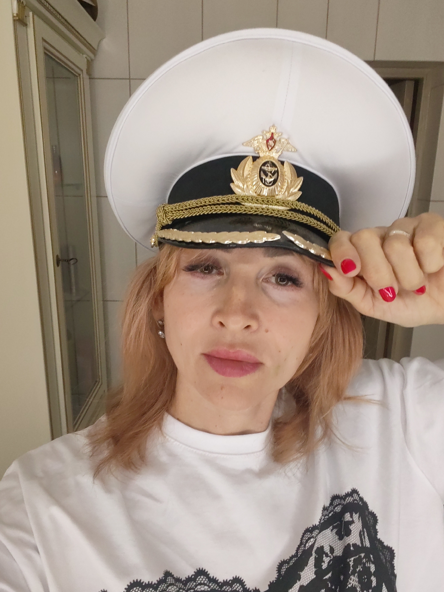 A woman with light brown hair and light skin is wearing a white uniform cap with gold trim and an emblem. She has red nail polish on her fingernails and is wearing a white t-shirt with a black lace design on it. She is holding the brim of the cap with her right hand. The background appears to be an indoor setting with tiled walls and a piece of furniture with glass doors. The image does not provide any information that would allow for its location to be specified as Bol'shie Mkhi, Russia.